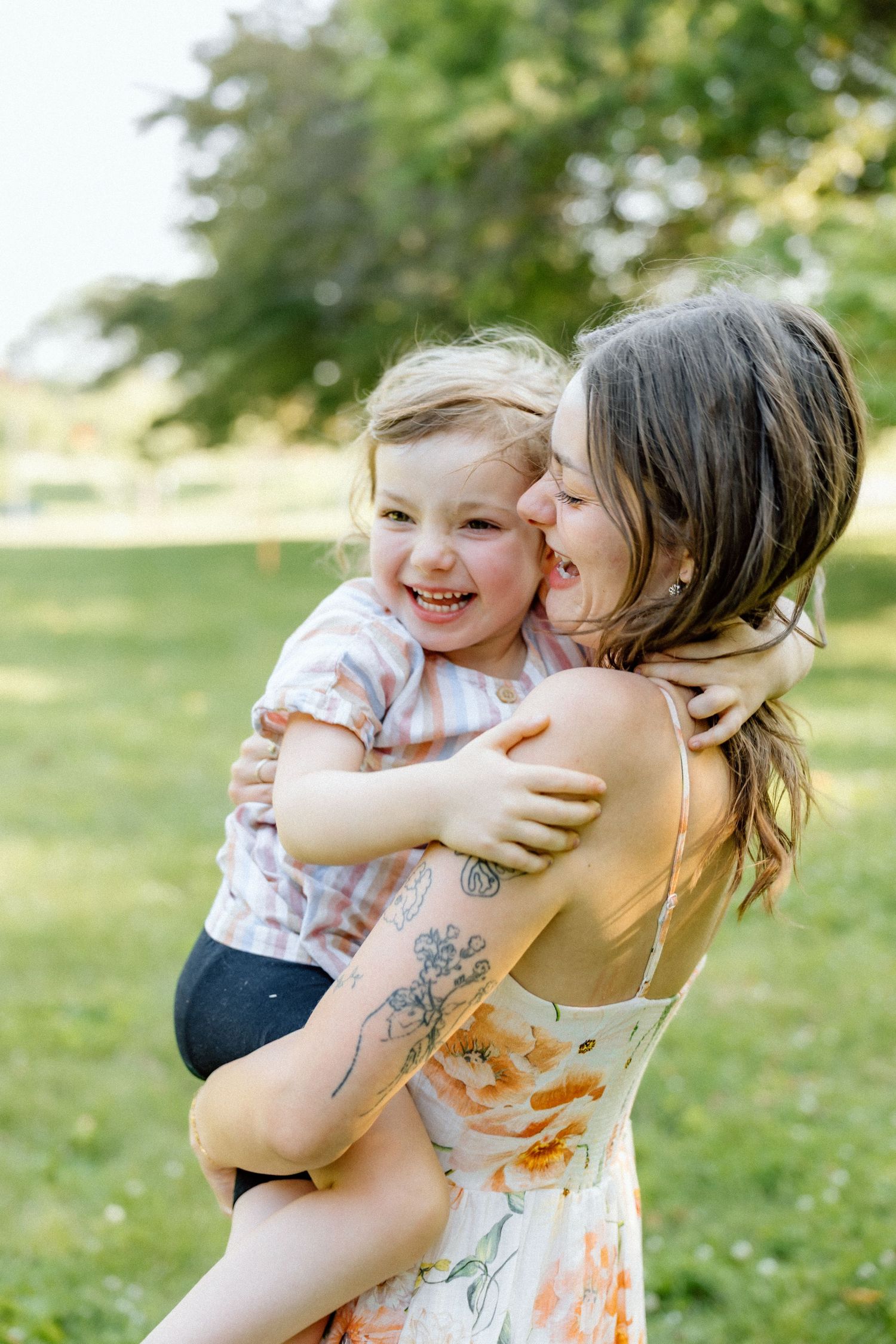 Mother kissing her smiling toddler daughter while holding her in a sunny green park.