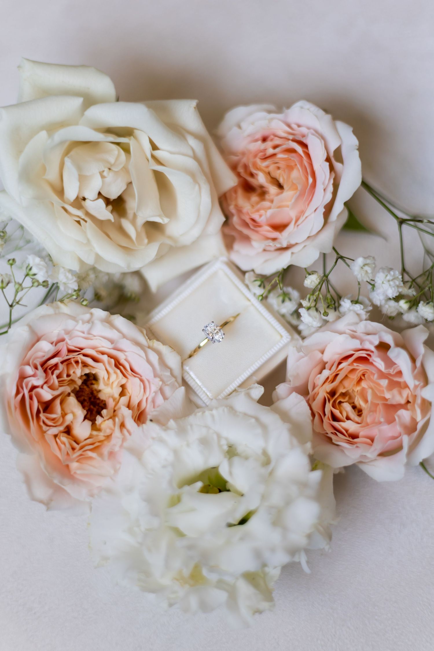 Elegant diamond engagement ring in cream box surrounded by blush pink and white roses and baby's breath.