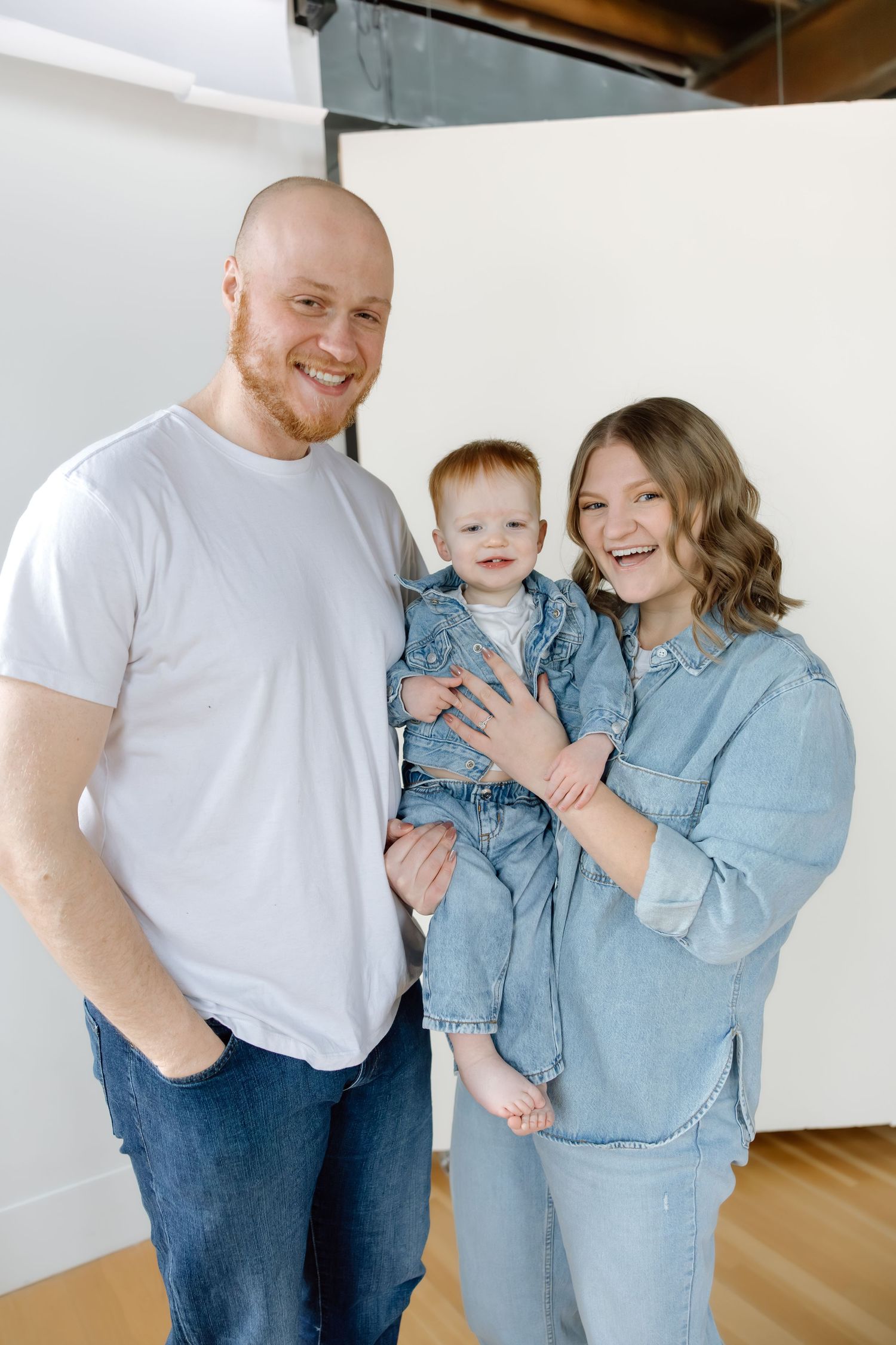 Happy family with redhead father, mother, and baby dressed in denim poses in a bright photo studio.