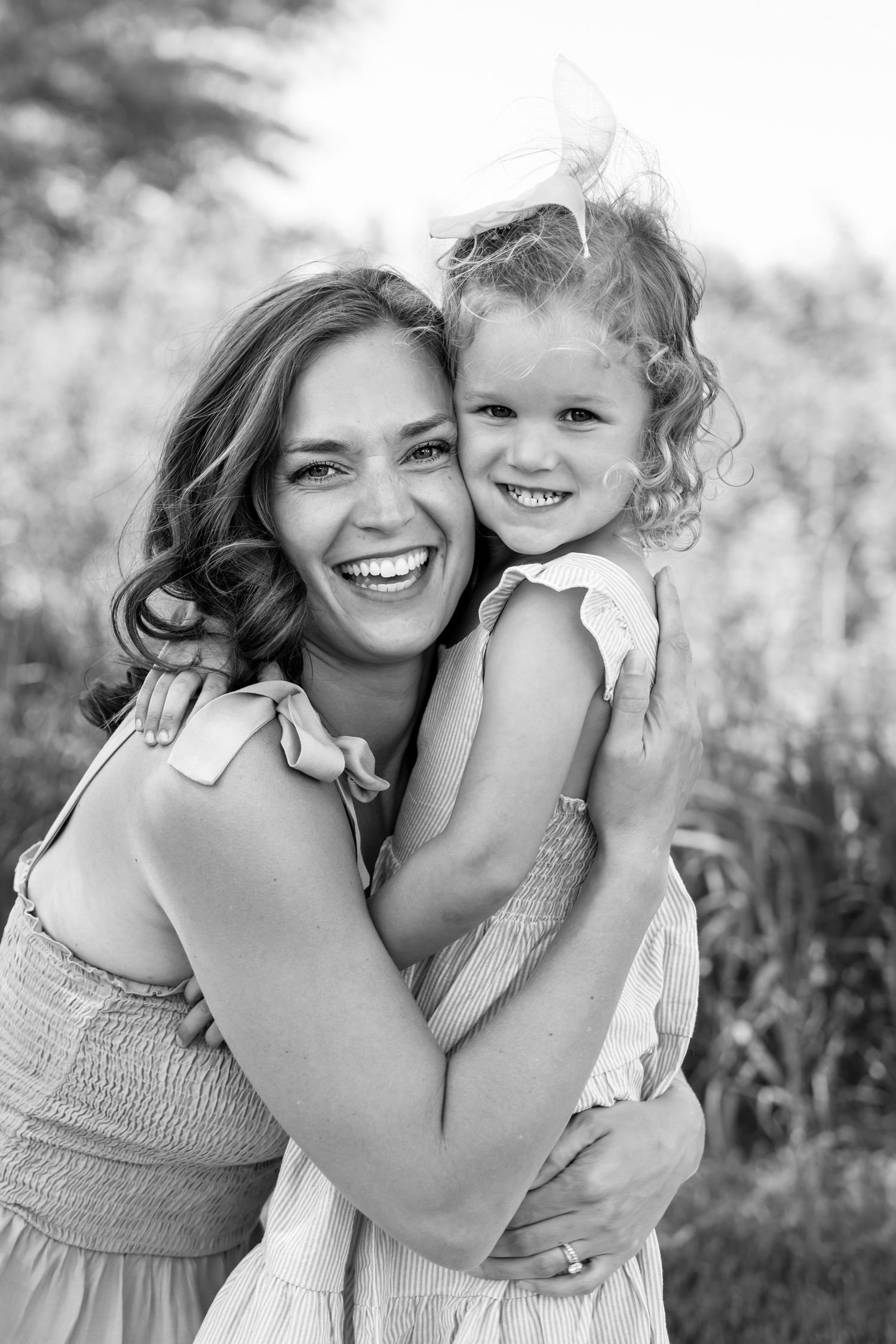 Black and white portrait of a laughing mother hugging her smiling young daughter outdoors.