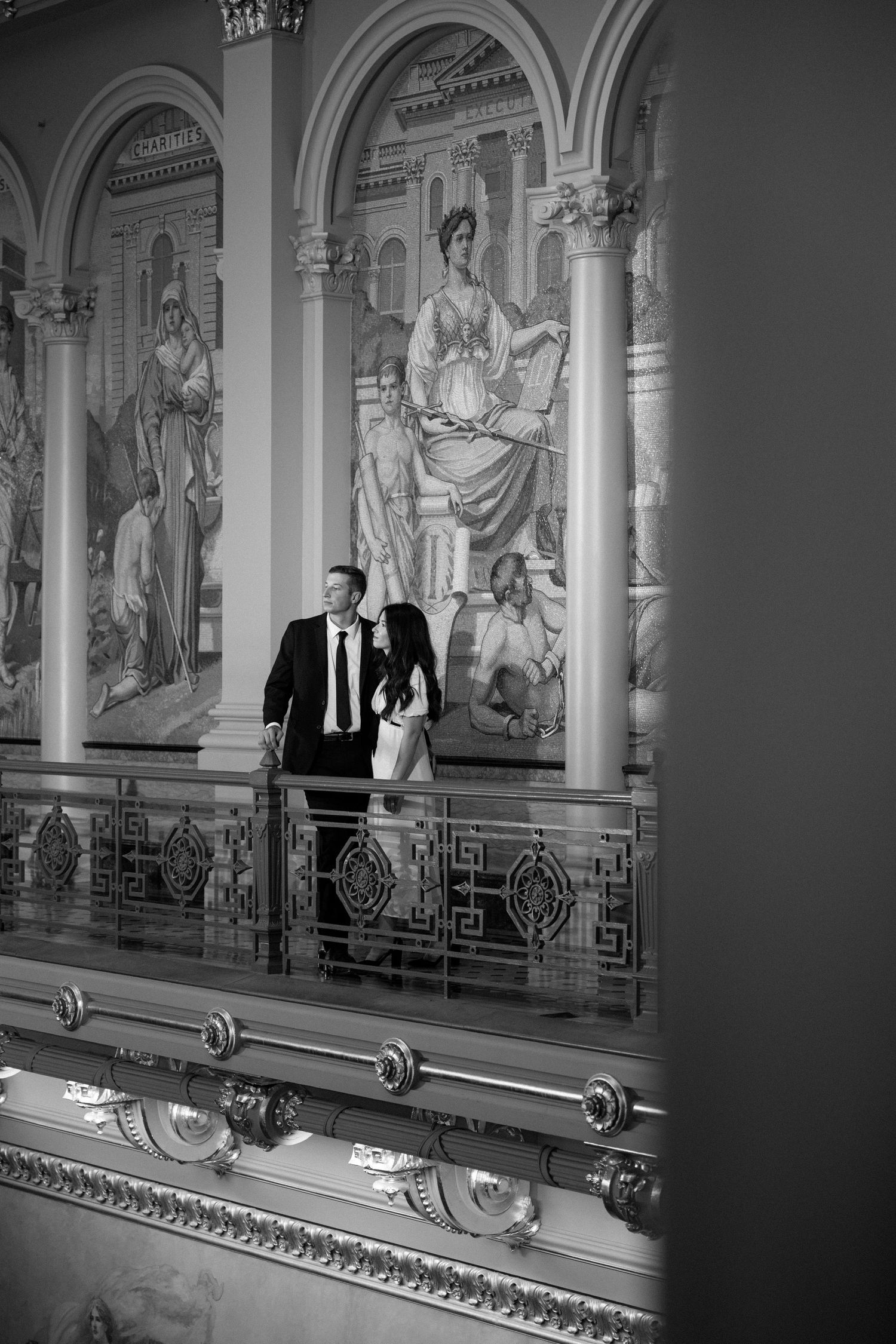 Black and white photo of couple walking a grand balcony adorned with classical fresco murals.