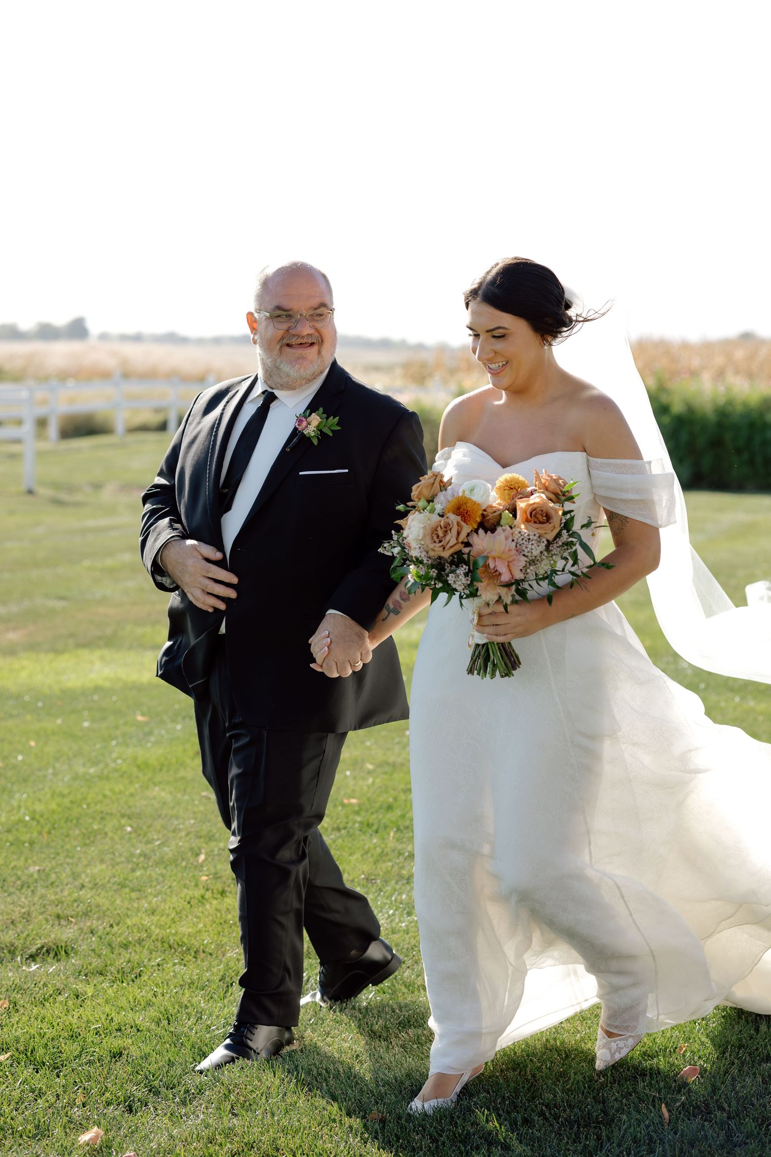 Smiling bride in off-shoulder gown holding peach bouquet walks with father across green lawn at outdoor wedding ceremony.