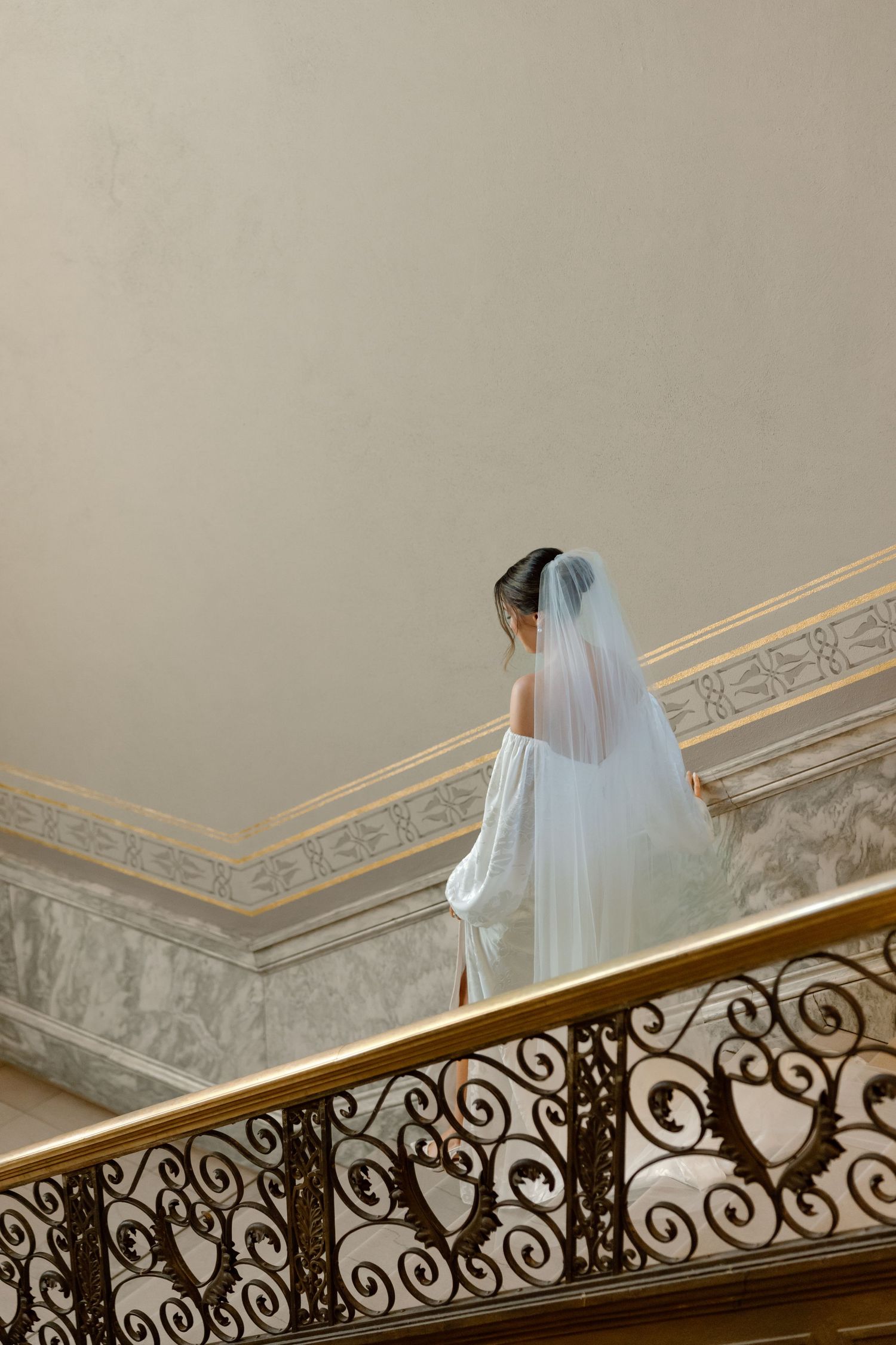 Bride in white gown and flowing veil descends ornate staircase with gold railing in elegant venue.