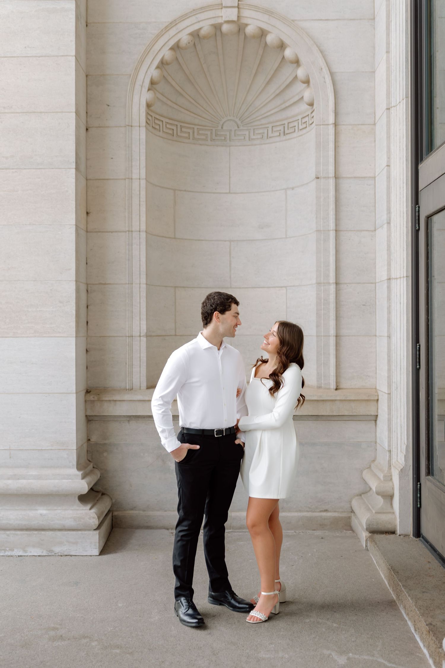 Elegant couple in white outfits standing close together beside ornate stone columns during an engagement photo session.