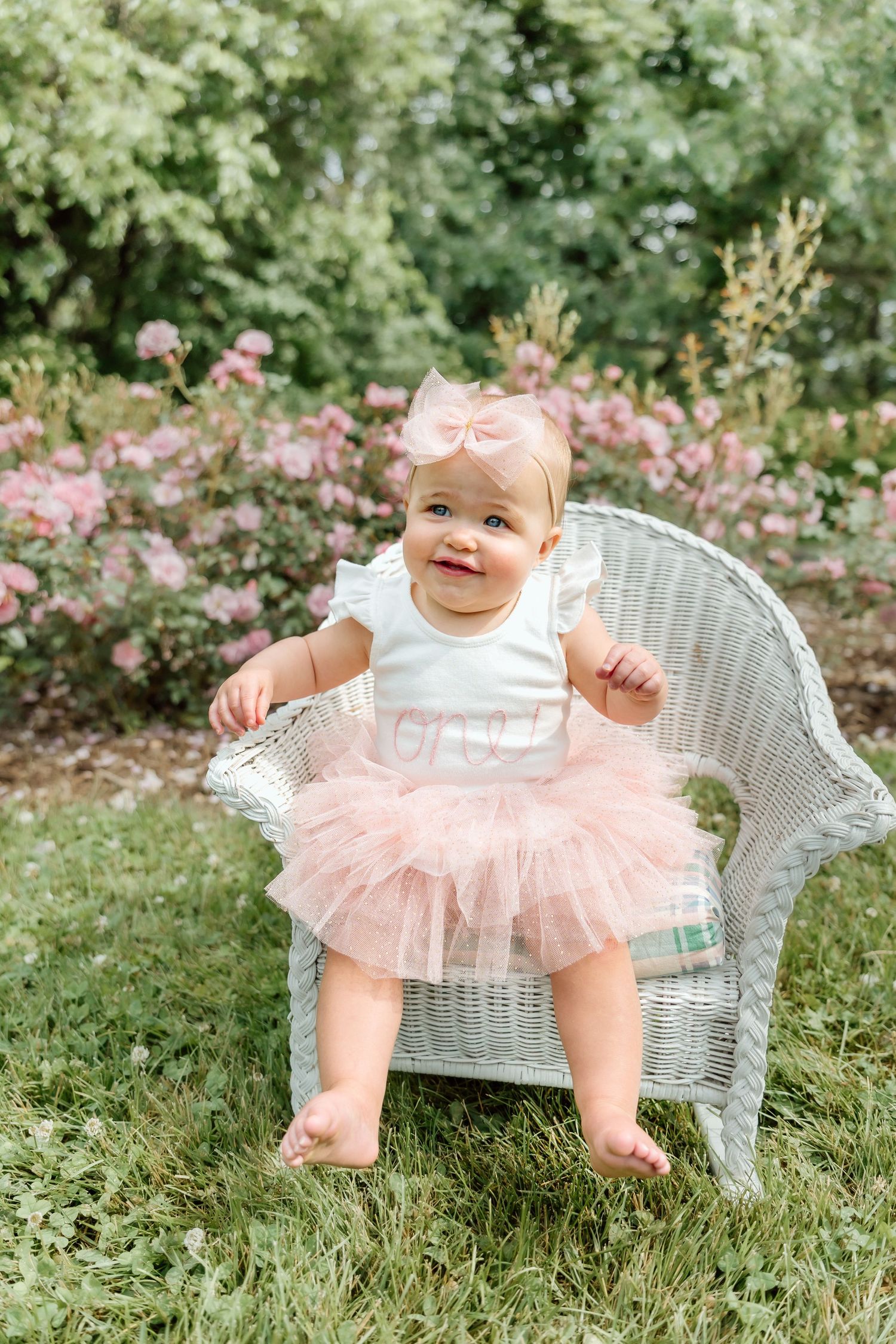 Adorable baby girl in pink tutu and bow headband standing in a wicker chair outdoors.