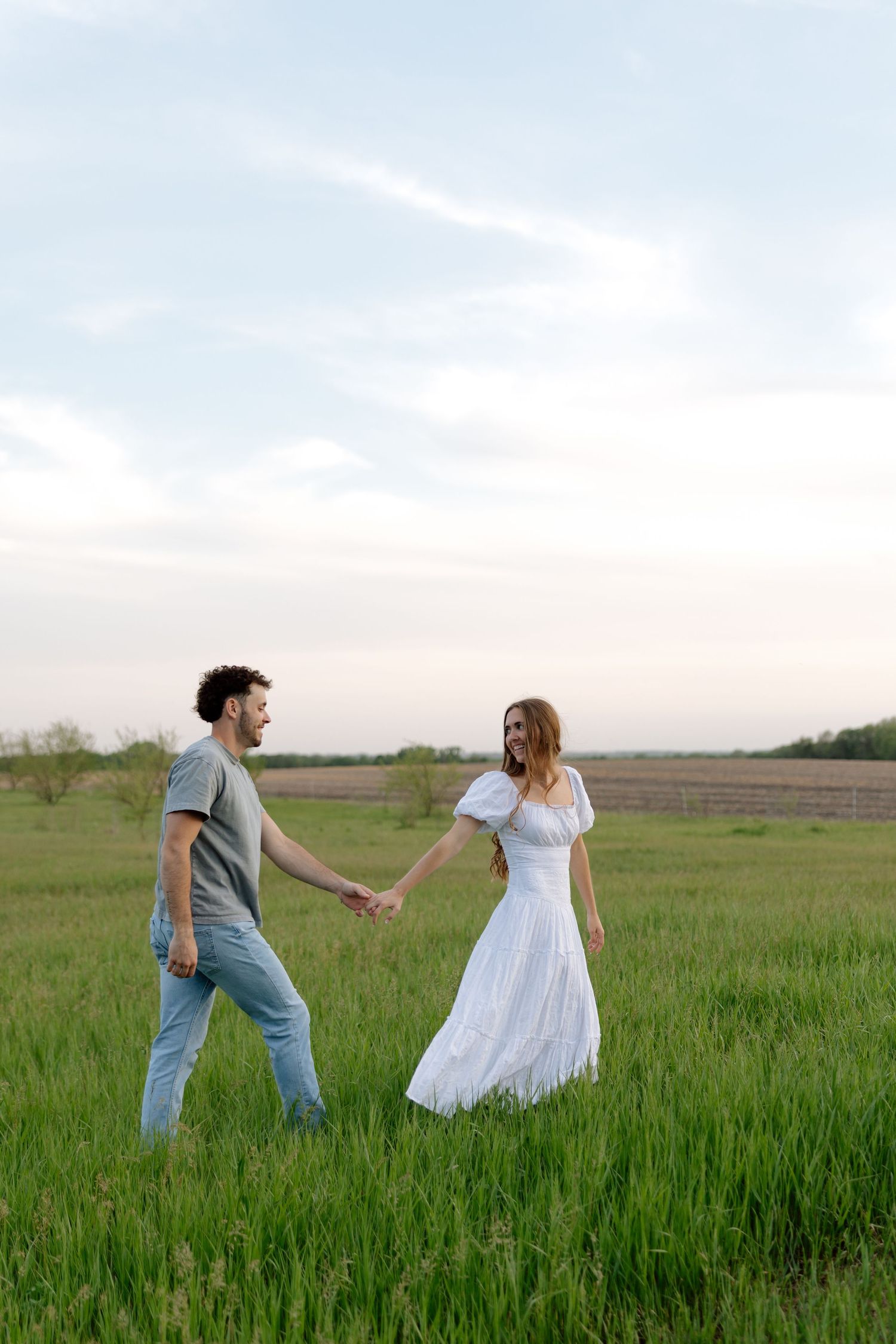 A couple holds hands walking through a lush green field, woman in white dress, man in gray shirt and jeans.