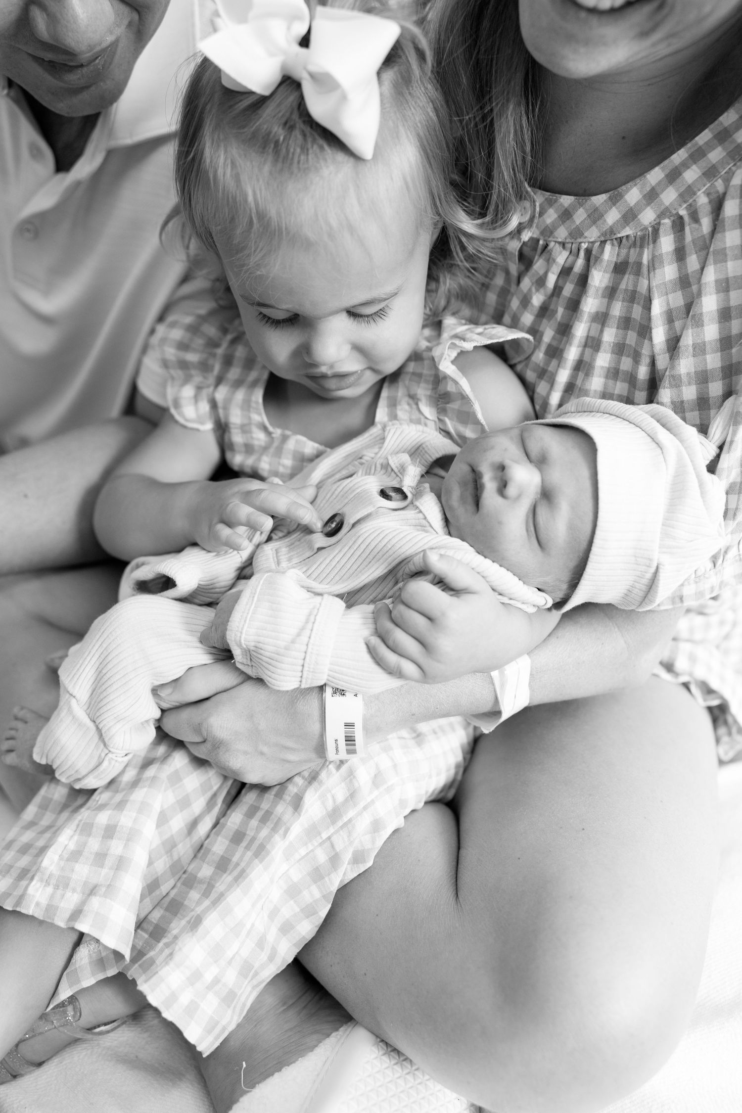 Black and white photo of a toddler girl with bow meeting her newborn sibling, held by their mother in a hospital setting.