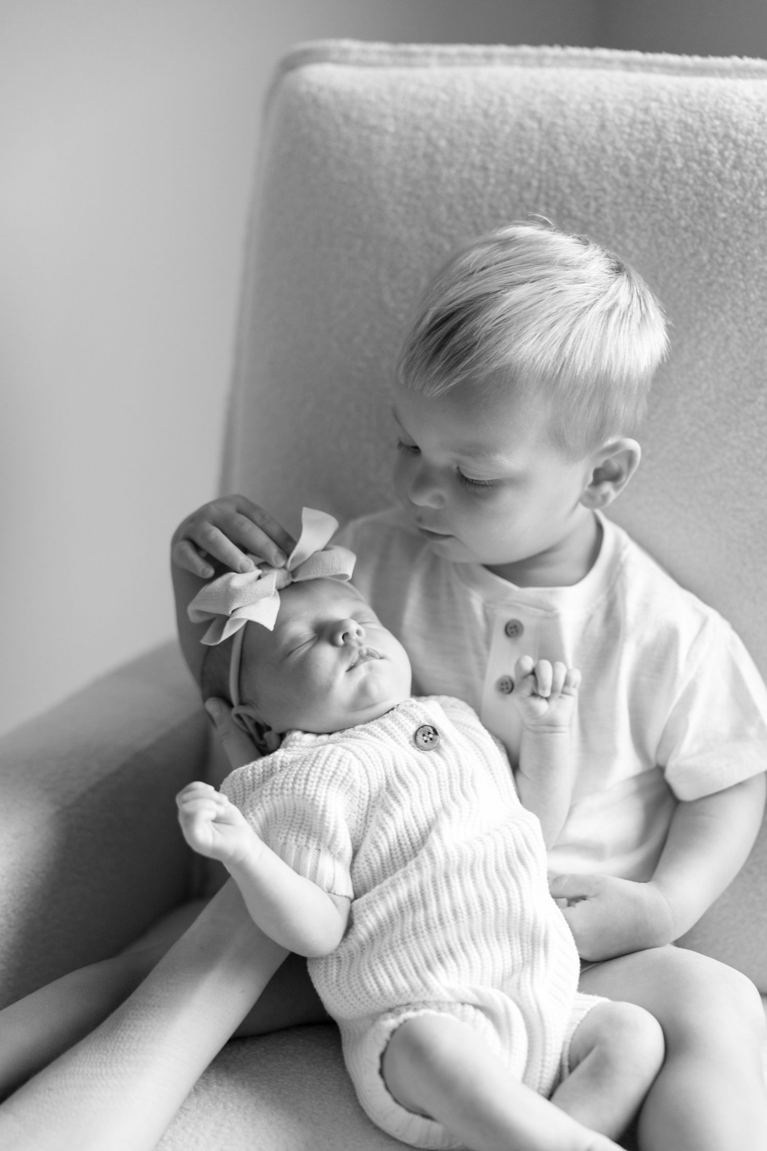 Black and white photo of a young boy kissing a sleeping newborn baby girl wearing a bow headband in a chair.