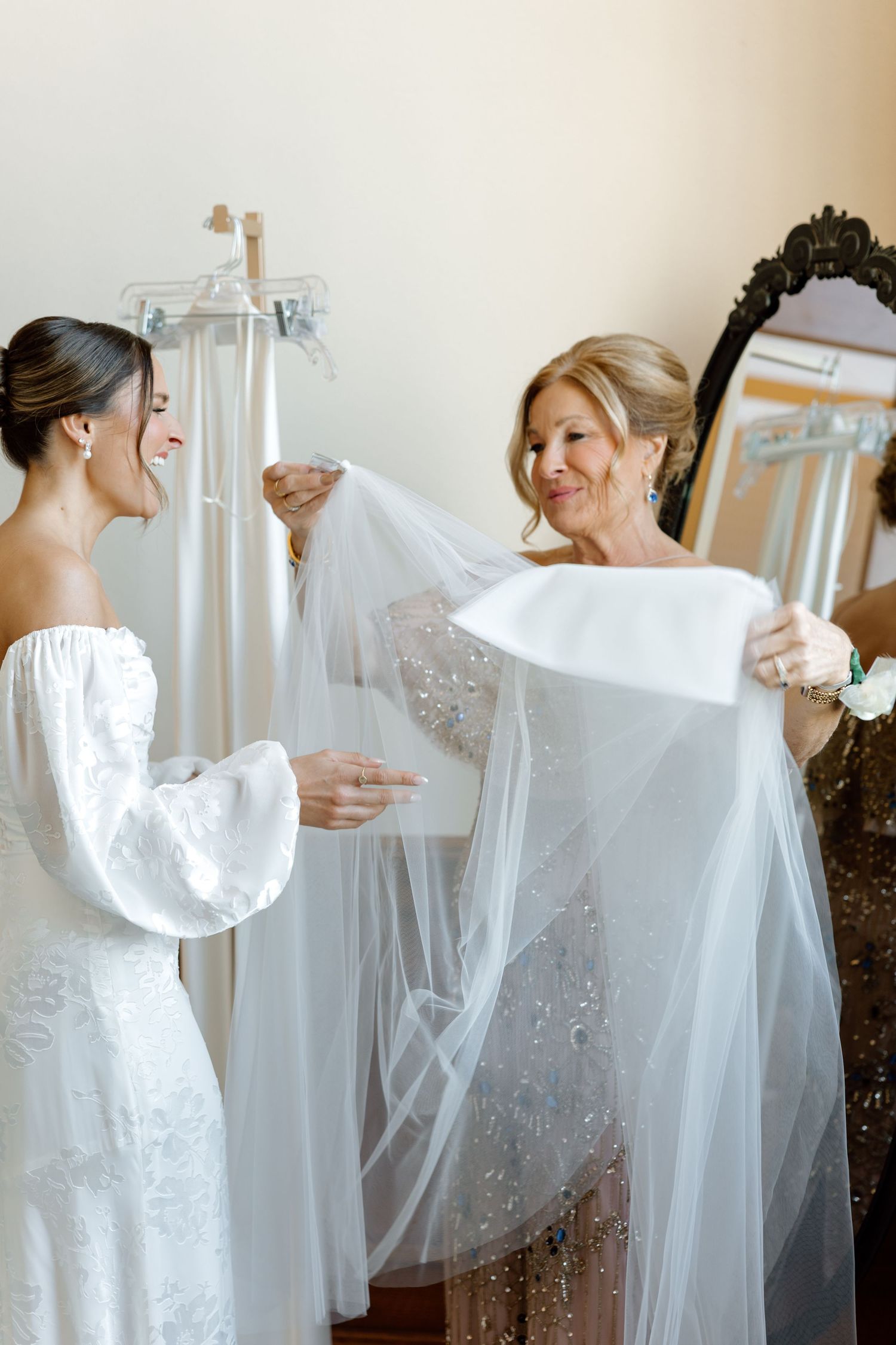Bride and older woman examining a white wedding veil together in a bright dressing room with gowns hanging nearby.