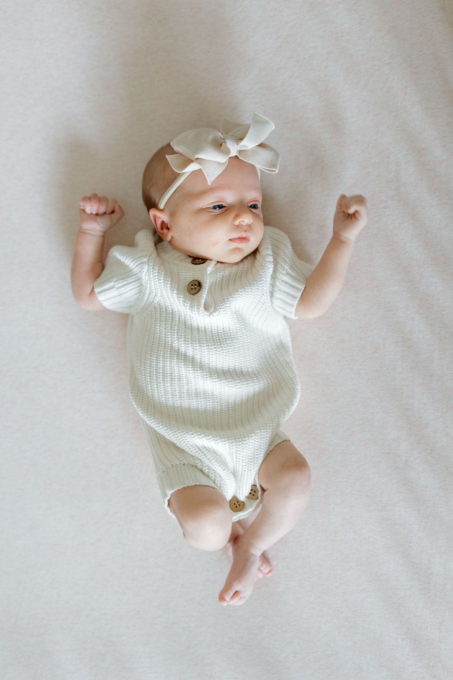 Newborn baby girl in a white knit romper and bow headband lying on her back with fists raised.