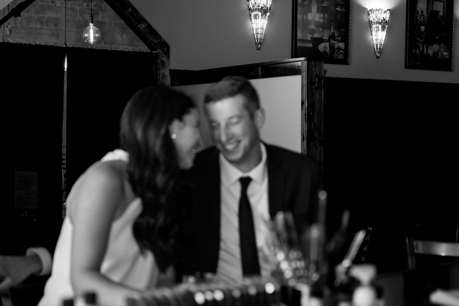 Black and white candid photo of laughing couple at a restaurant table, woman whispering to man in tie.