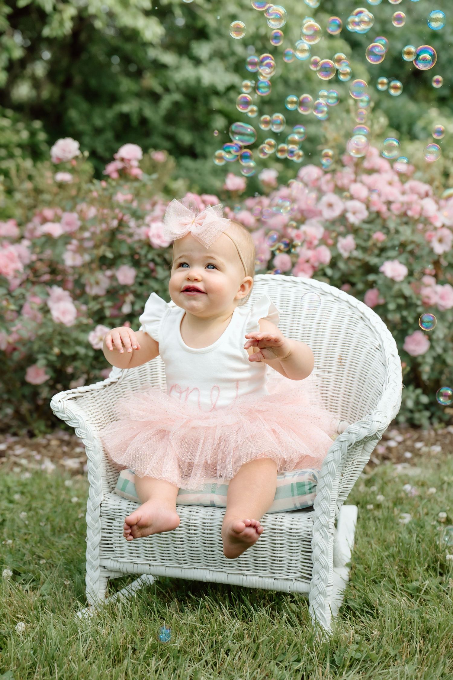 Happy baby girl in pink tutu sitting in wicker chair surrounded by floating soap bubbles.