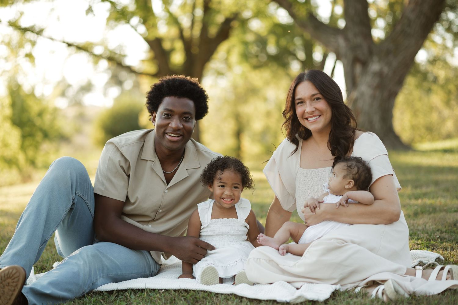 Happy interracial family of four sitting together on a blanket in a sunny park setting.