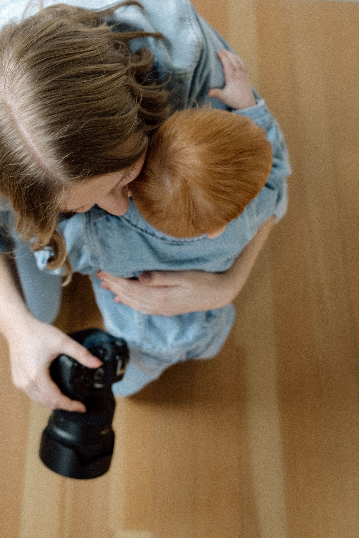 Overhead view of a photographer in denim hugging a redheaded toddler while holding a camera.