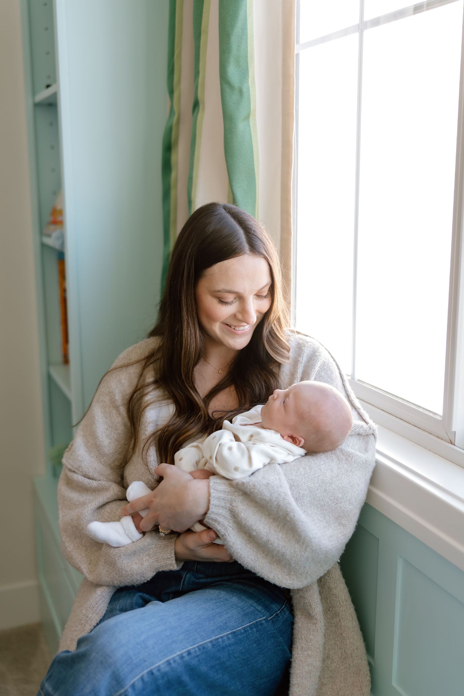 Smiling mother holding newborn baby by a bright window in a nursery.