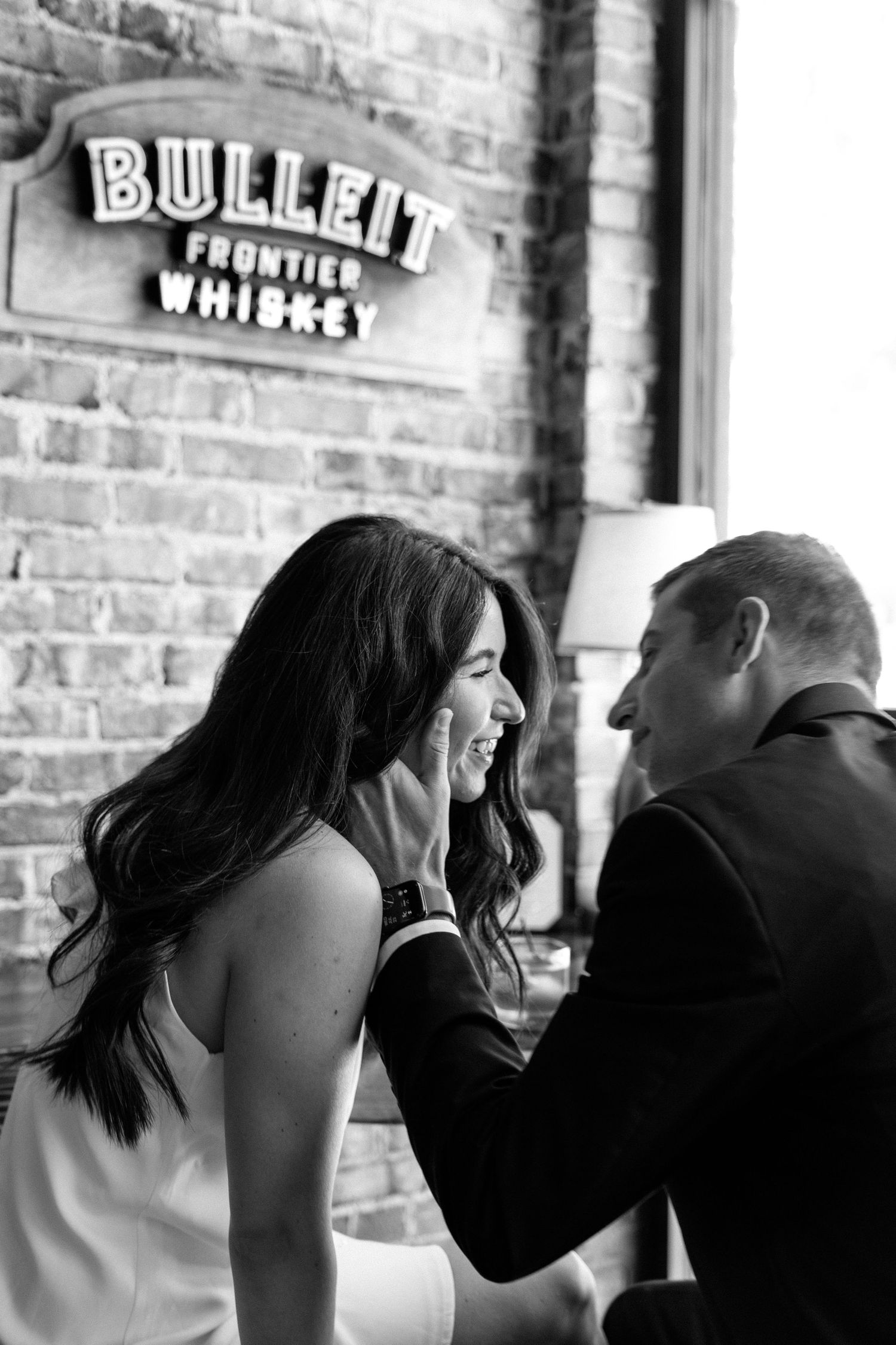 Black and white photo of a couple laughing together in front of a Bulleit Frontier Whiskey neon sign on brick wall.