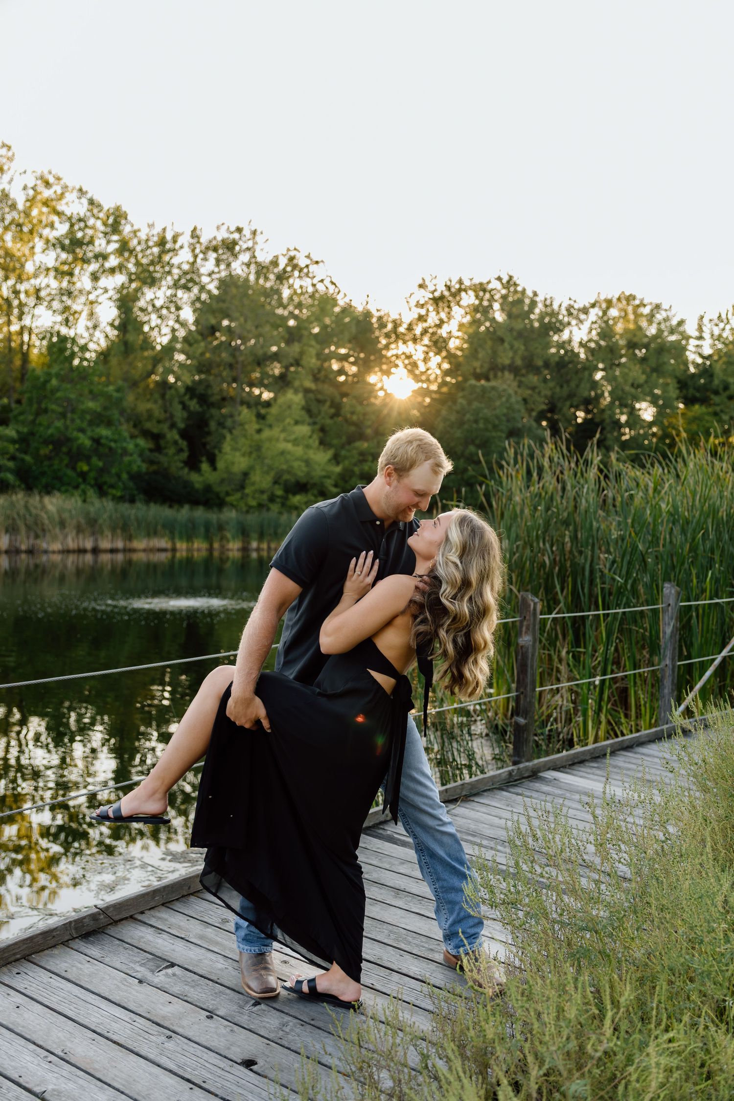 Couple sharing a romantic dip pose on a wooden dock at sunset, surrounded by a serene lake and lush greenery.