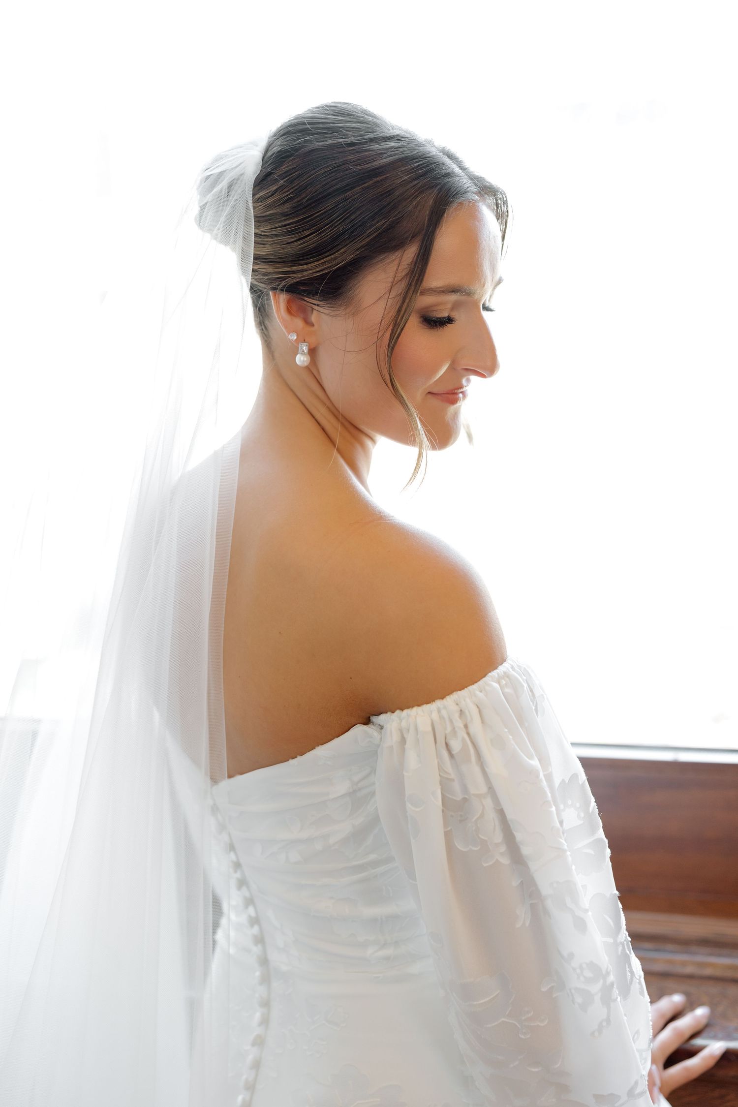 Elegant bride in off-shoulder lace wedding gown with veil and pearl earrings smiling by bright window.