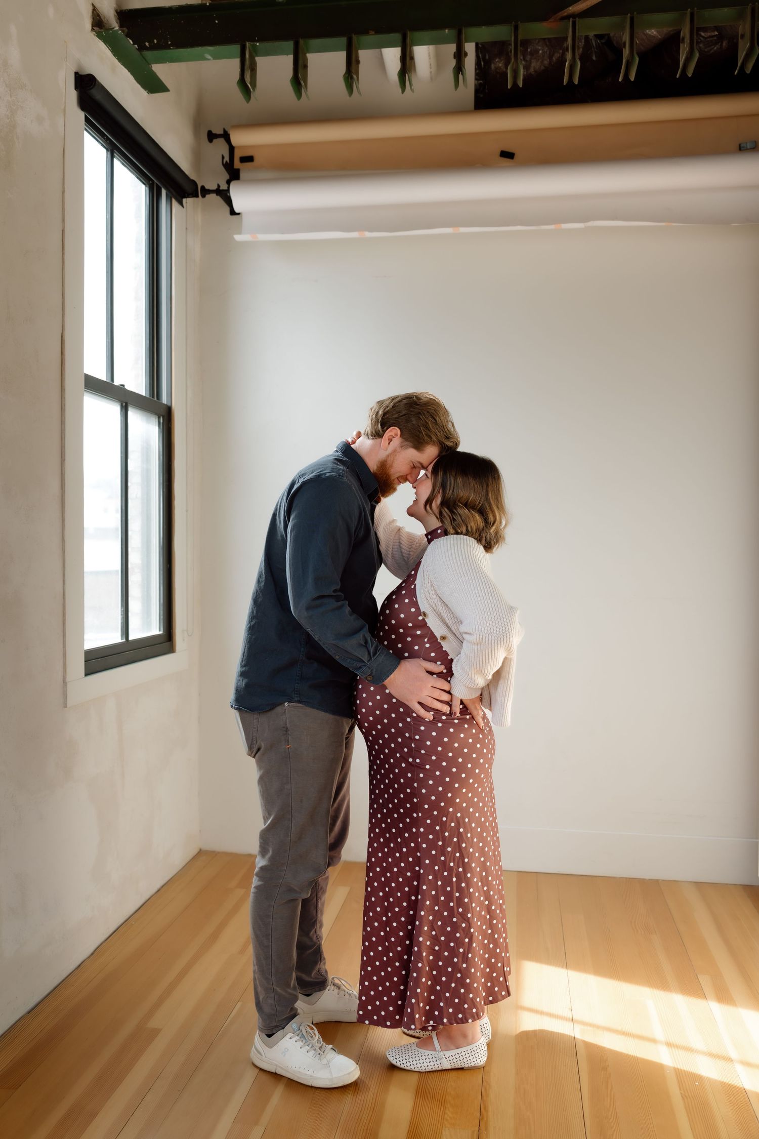 A couple shares a romantic forehead touch while the pregnant woman's baby bump is visible in a polka dot dress.