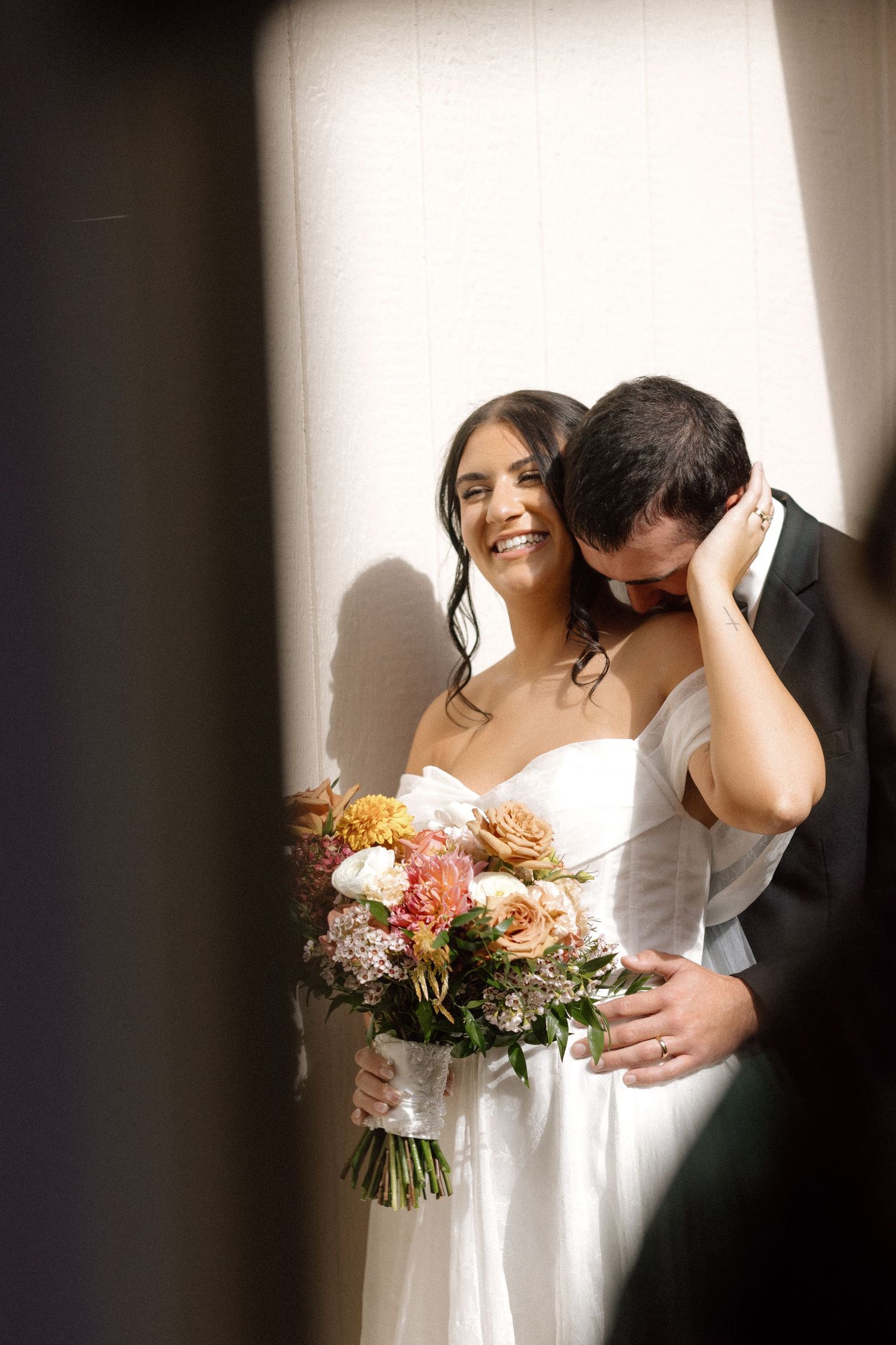 Smiling bride holding colorful bouquet as groom kisses her cheek against white wall.
