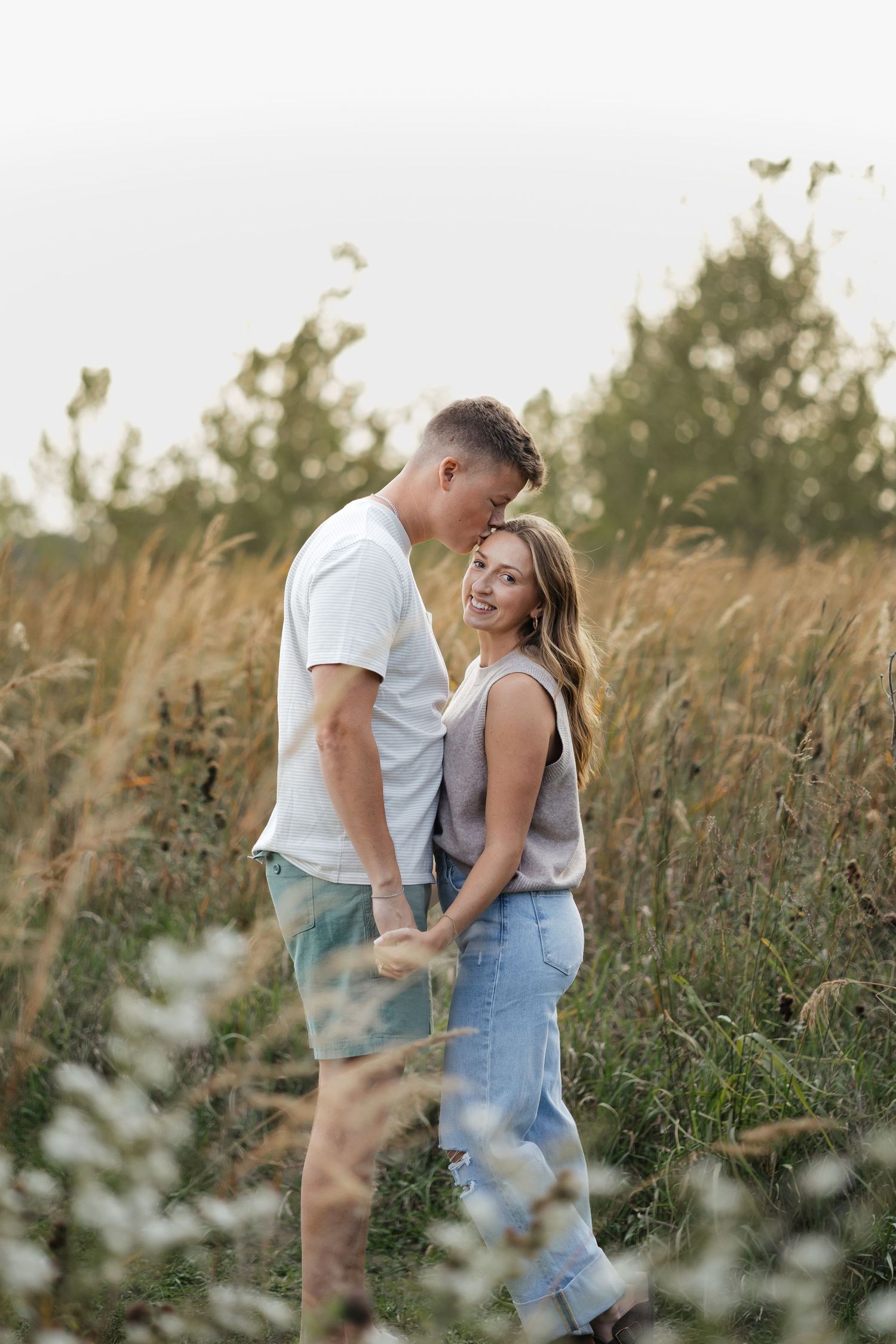 Couple holding hands in tall grass field at sunset, woman smiling at camera.
