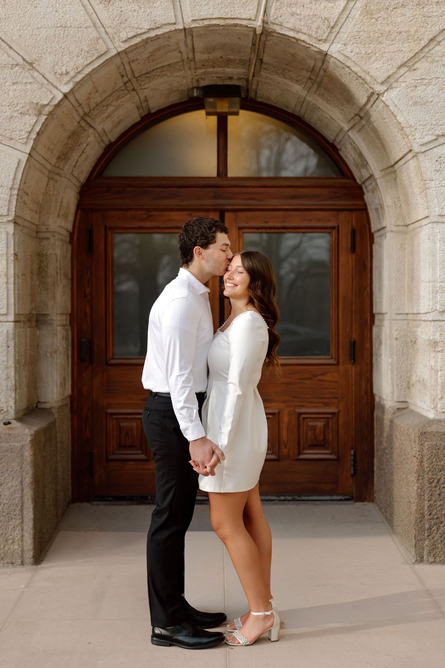 Couple shares a tender kiss in front of a large wooden arched doorway of a stone building.