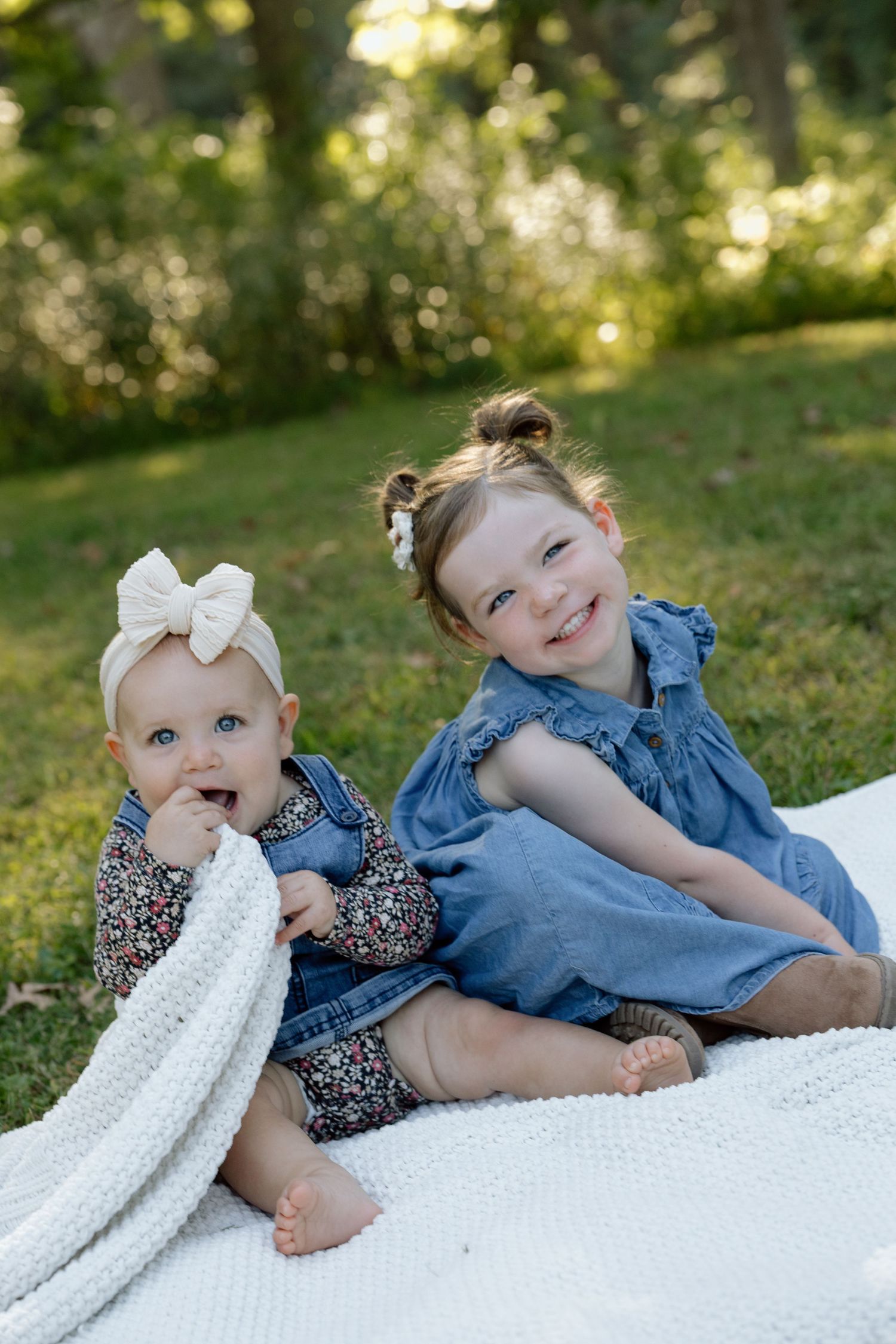 Two young sisters sitting on a white blanket outdoors, smiling happily in a green park setting.