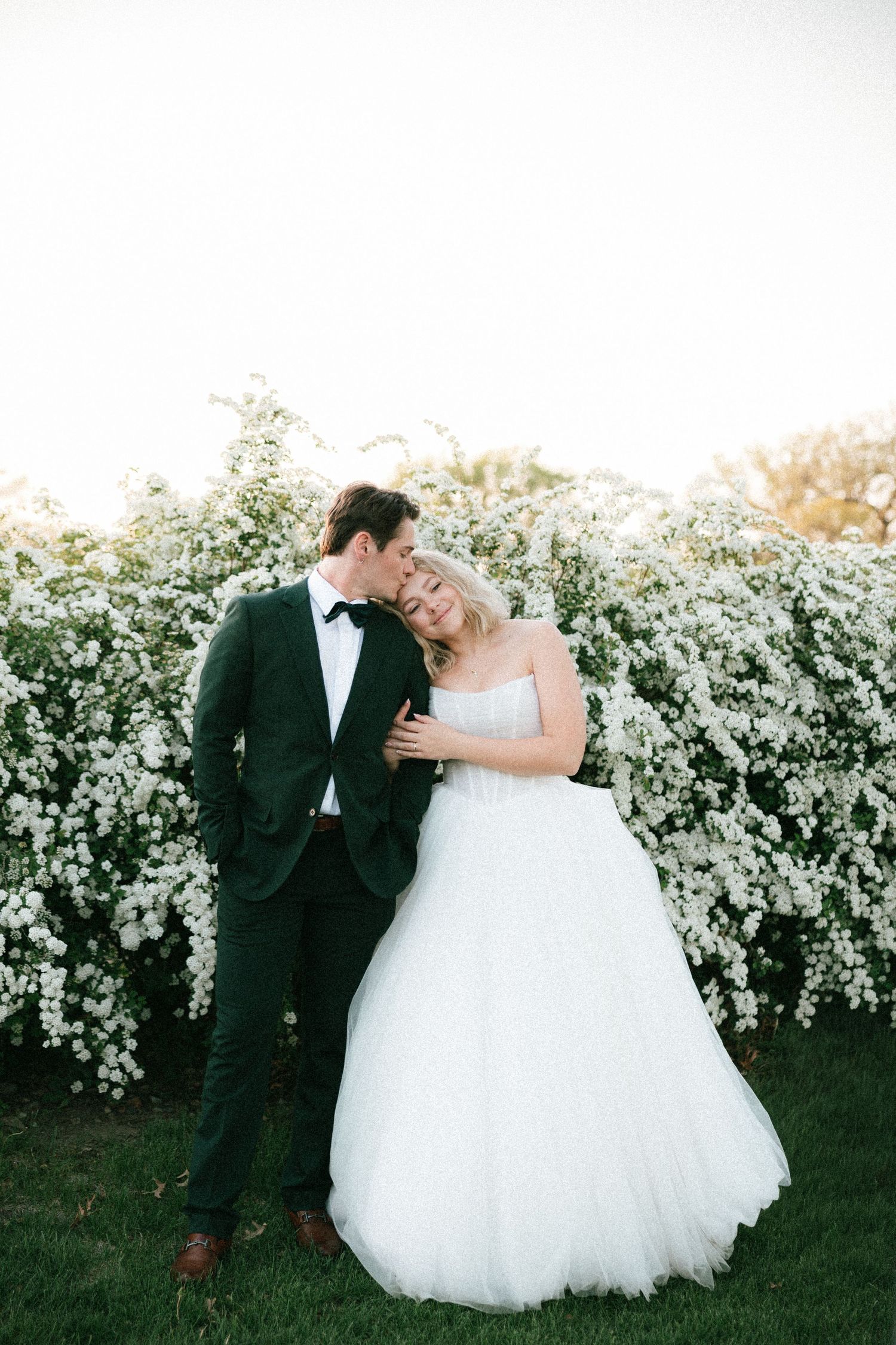 A bride and groom share a tender moment in front of white blooming flowers, she in a strapless tulle gown, he in a dark tuxedo.