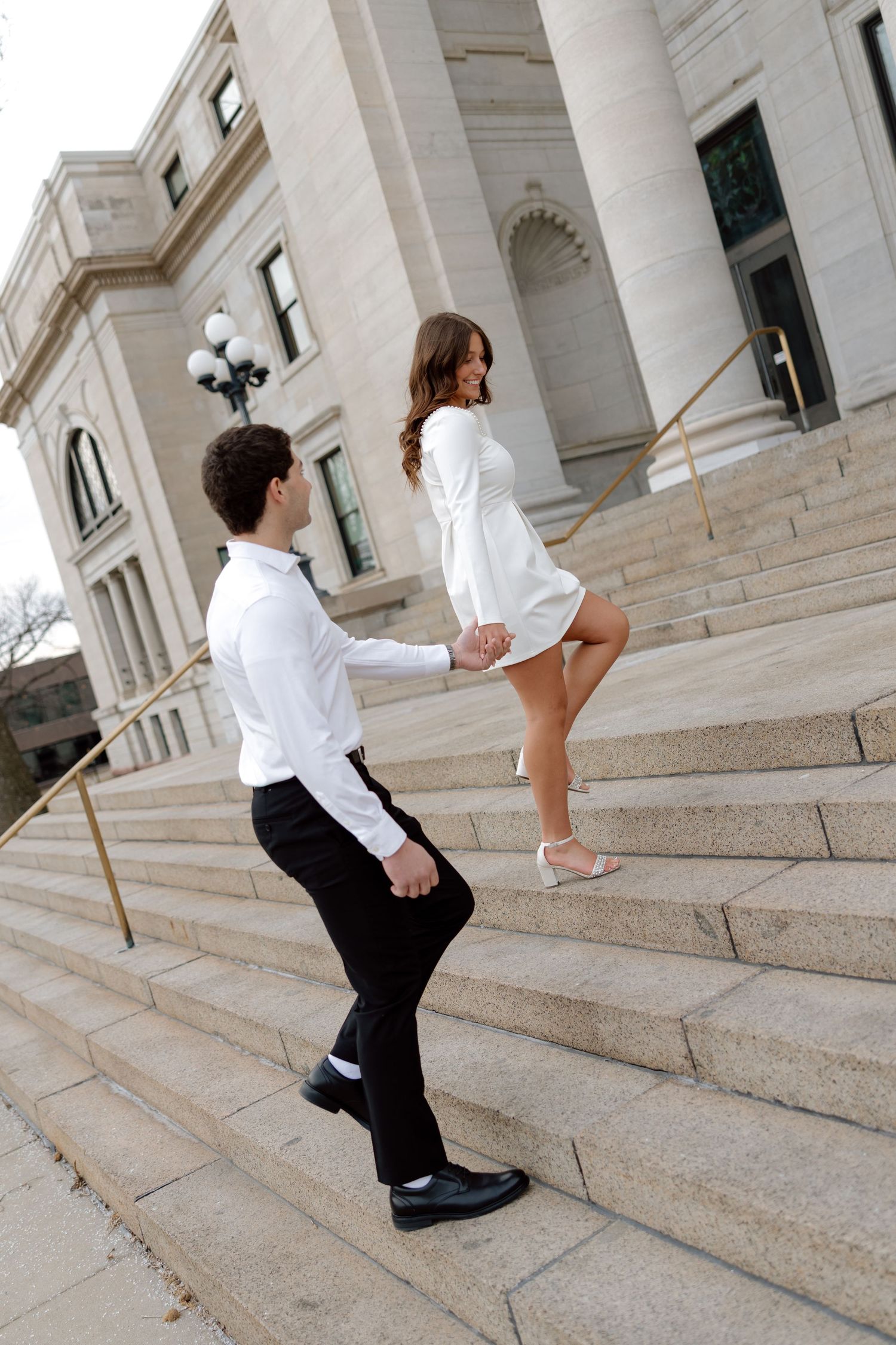 Couple in white outfits holding hands on grand stone building steps, woman leading man upward in elegant attire.