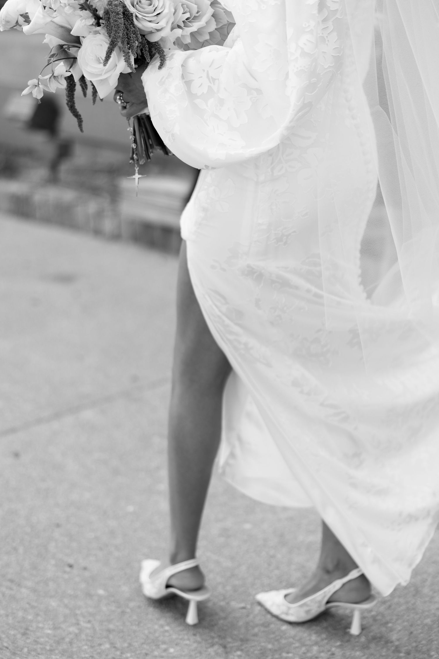 Black and white close-up of bride in lace gown and heels holding floral bouquet outdoors.