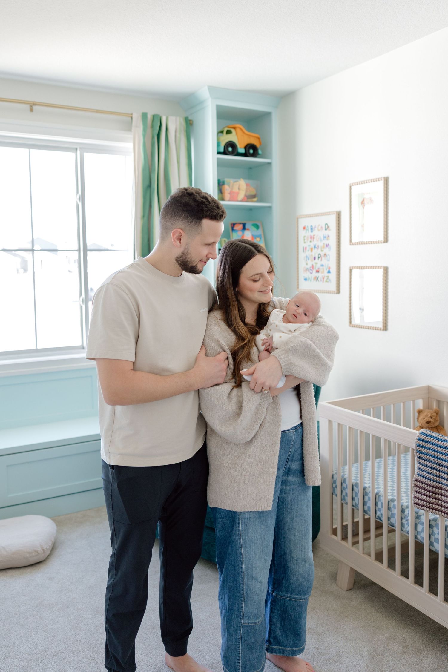 New parents stand together in a light blue nursery, lovingly gazing at their newborn baby near a white crib.