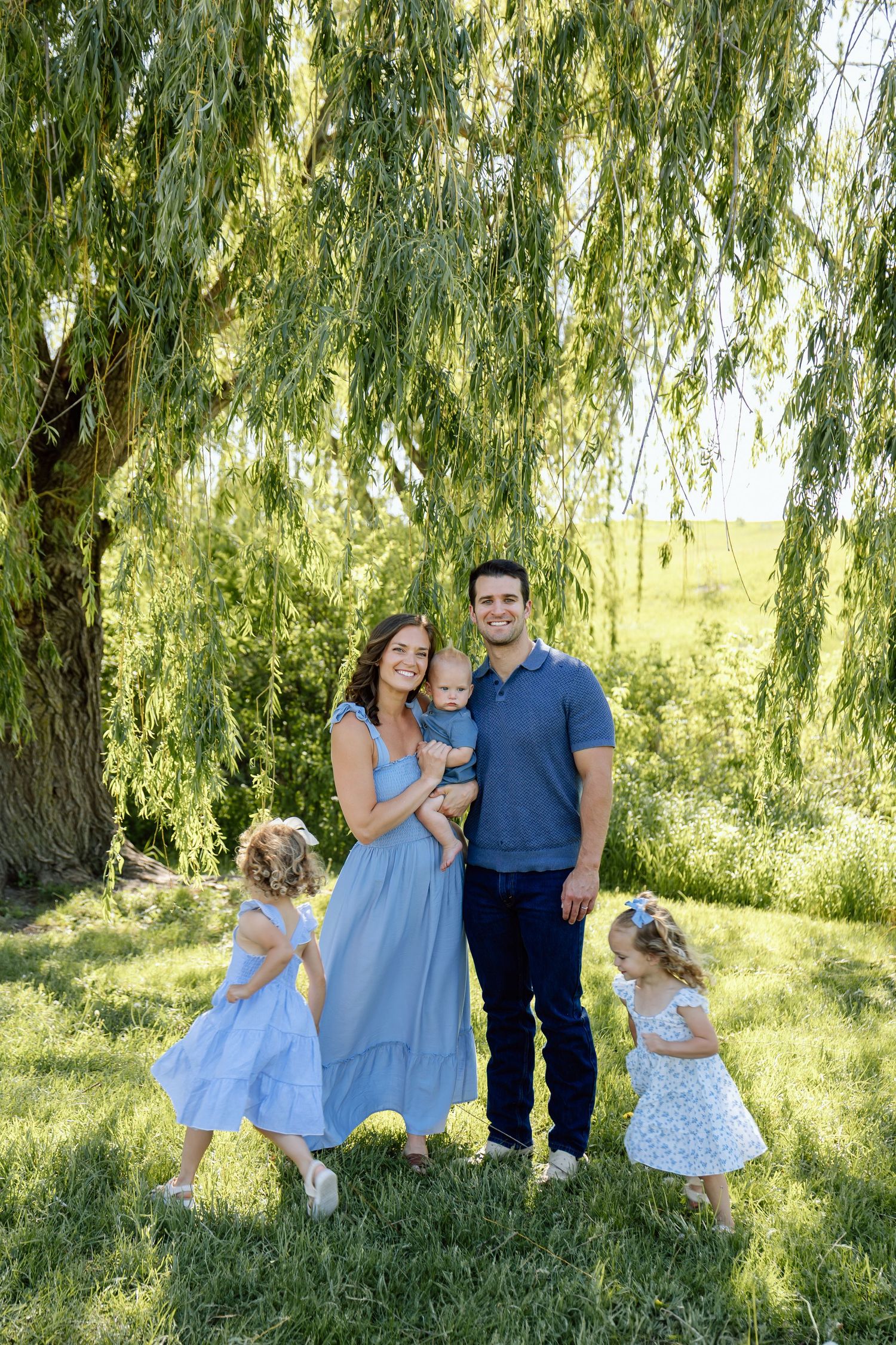 Family of five posing under a weeping willow tree in coordinating blue outfits on a sunny day.