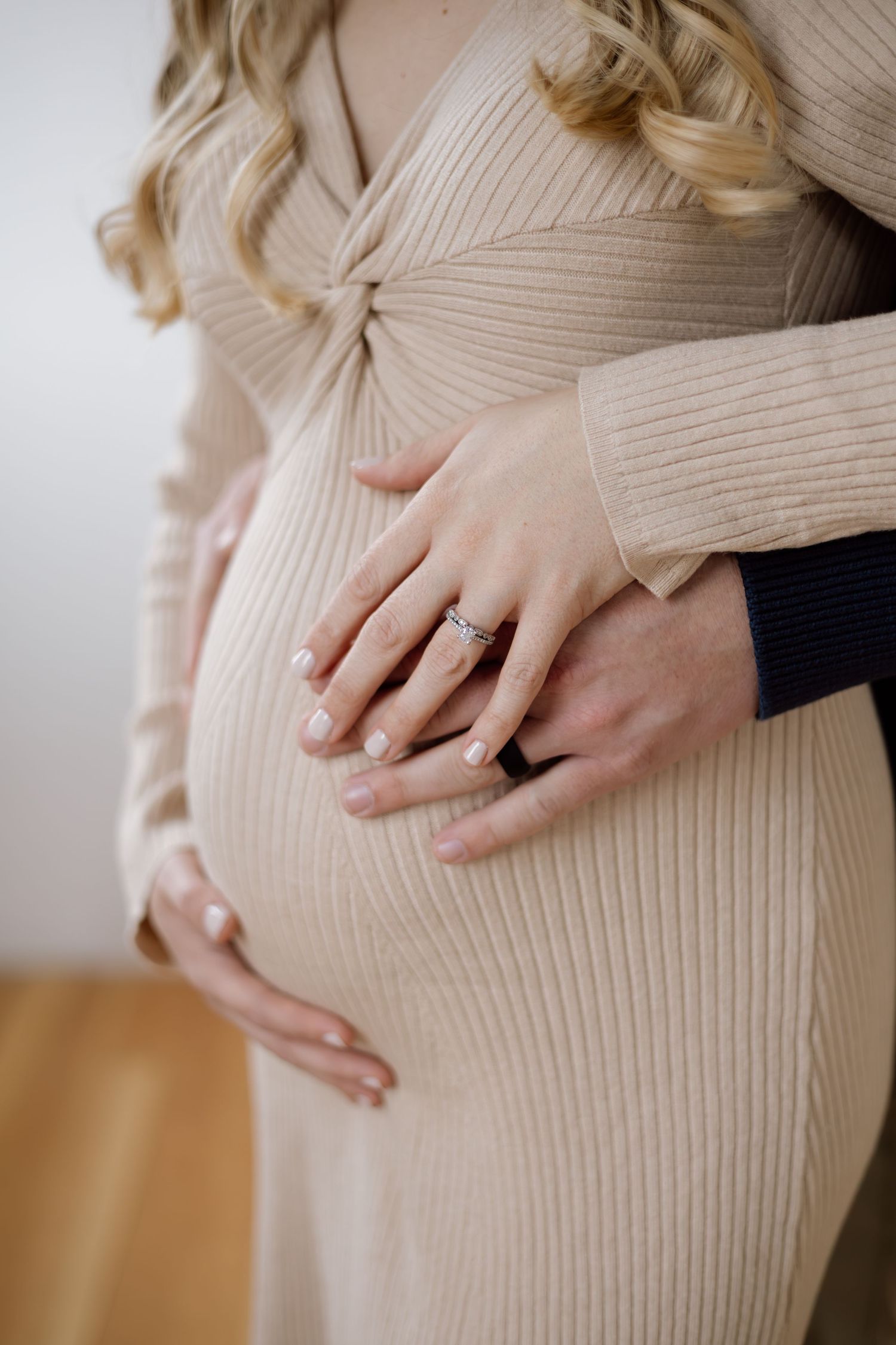 Close-up of a pregnant woman in a beige ribbed dress with her partner's hands resting on her baby bump.