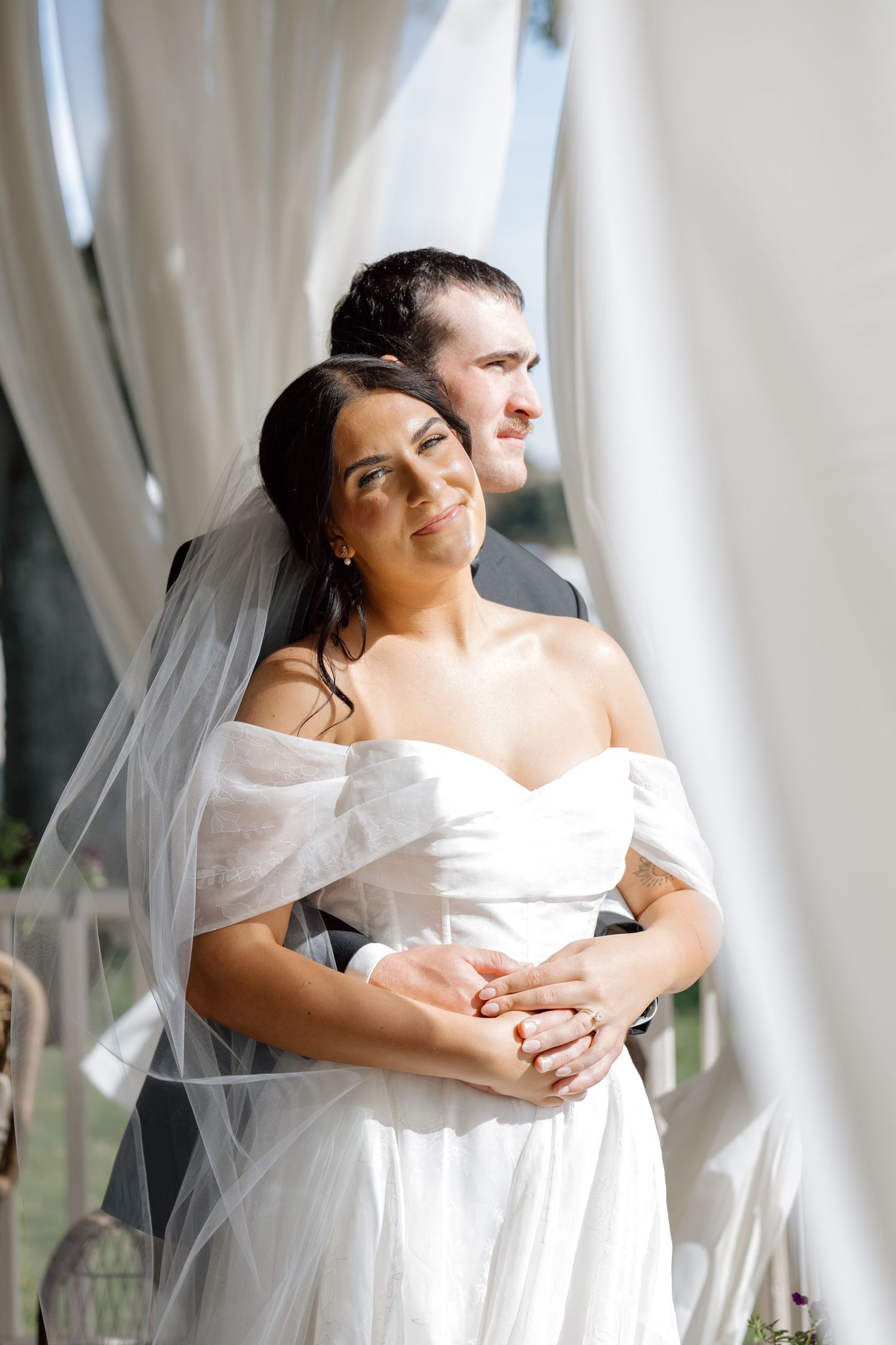 Bride in off-shoulder white gown with veil embraced by groom surrounded by flowing white curtains outdoors.