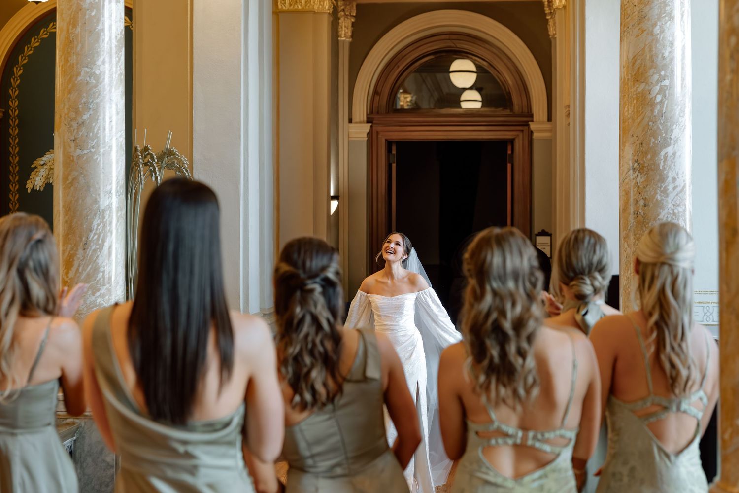 Bride in white gown and veil reveals herself to bridesmaids in sage dresses in an elegant columned hall.