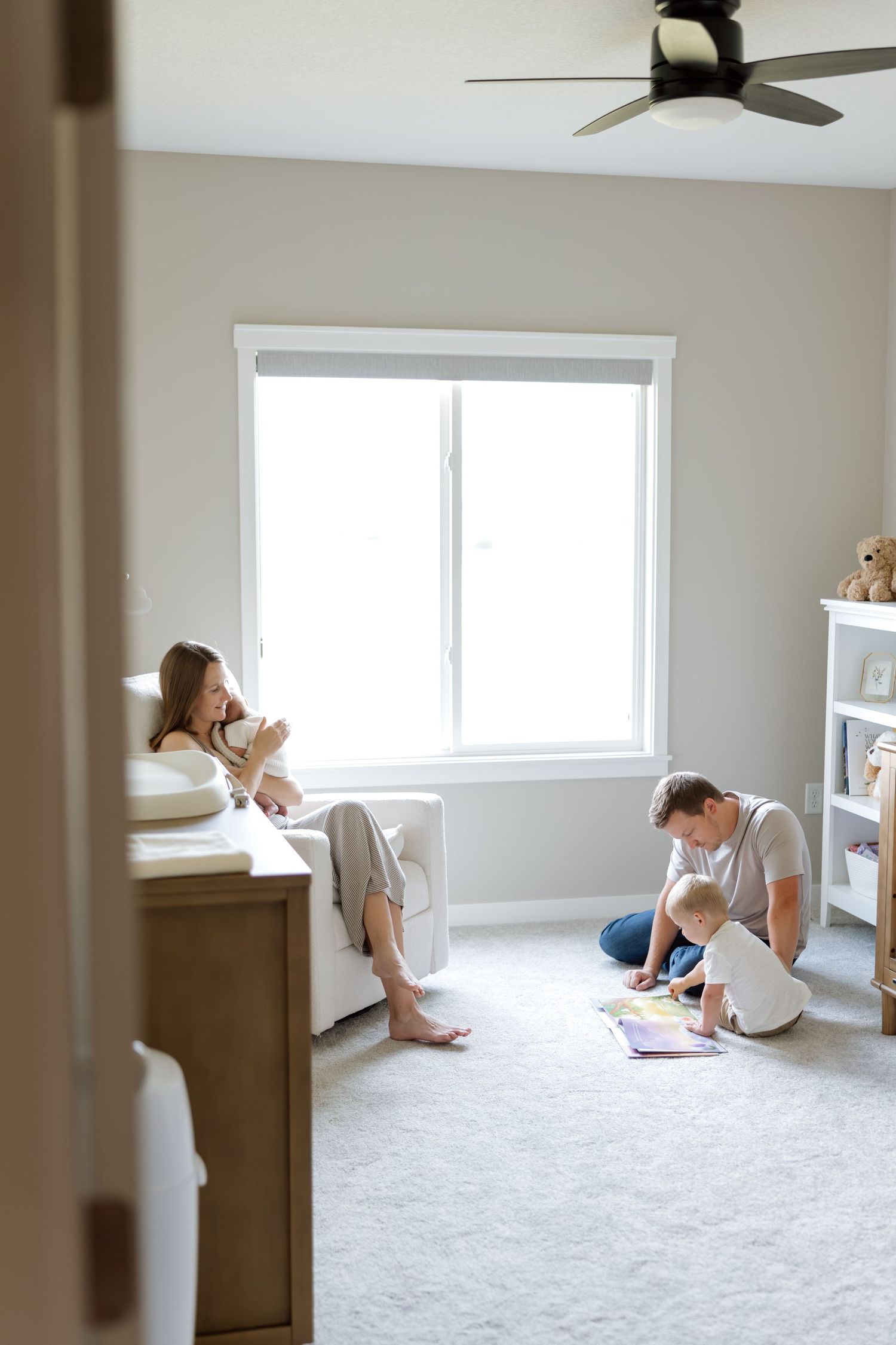 Family relaxing in a bright nursery, mother holding newborn while father reads with toddler on carpet.