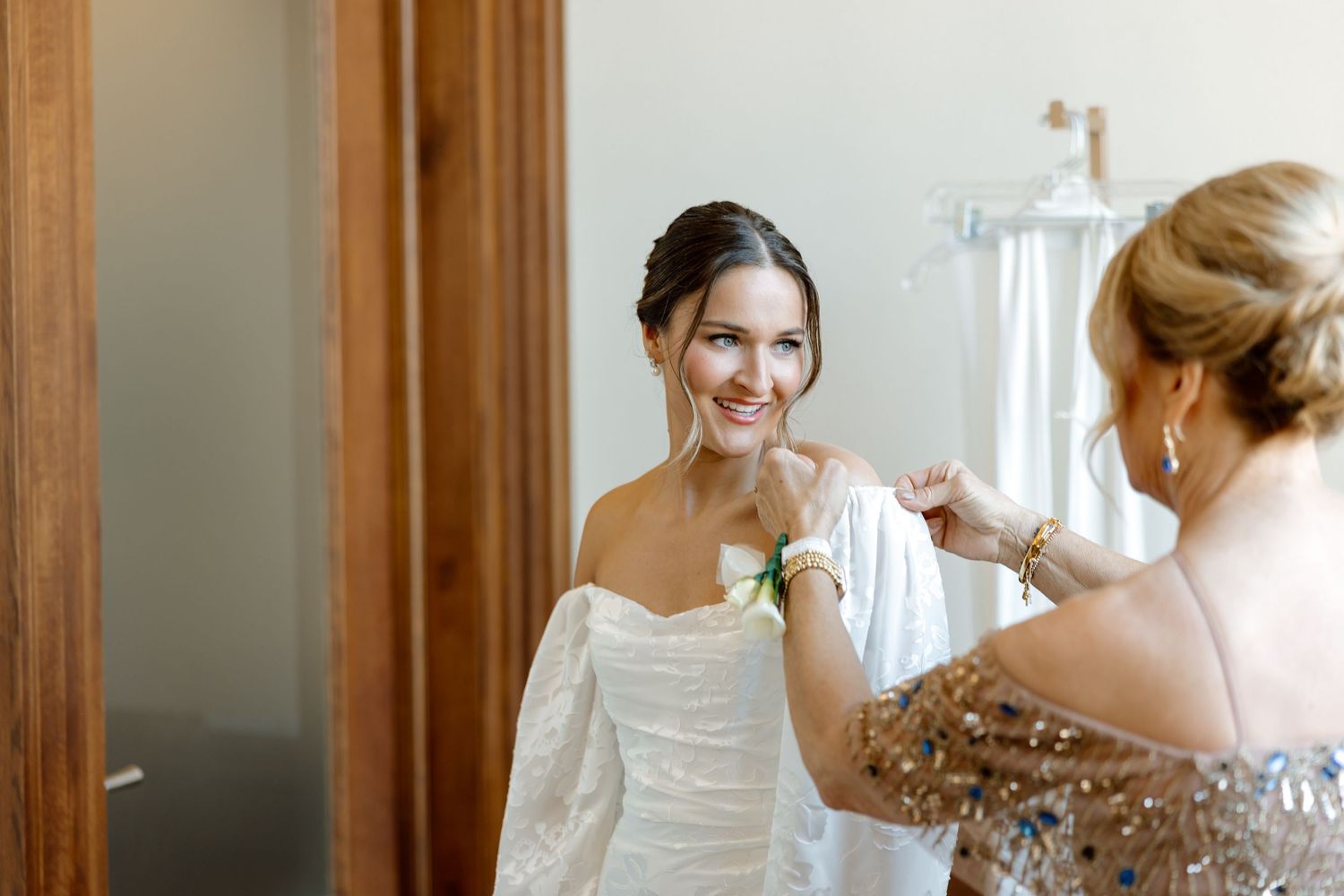 Bride smiling as bridesmaid helps fasten her white off-shoulder wedding gown in a bright dressing room.