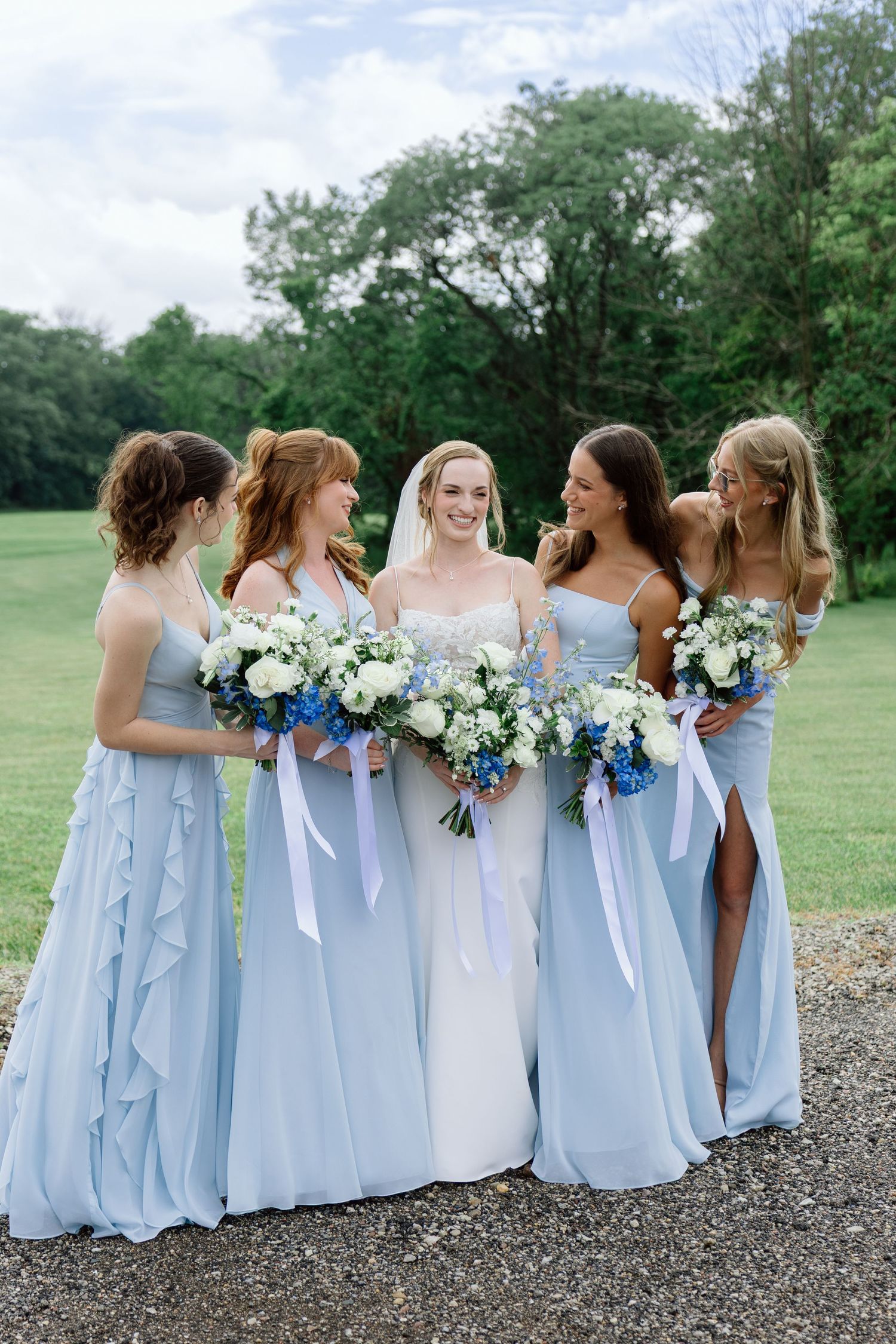Bride in white gown stands with four bridesmaids in light blue dresses holding white and blue bouquets outdoors.