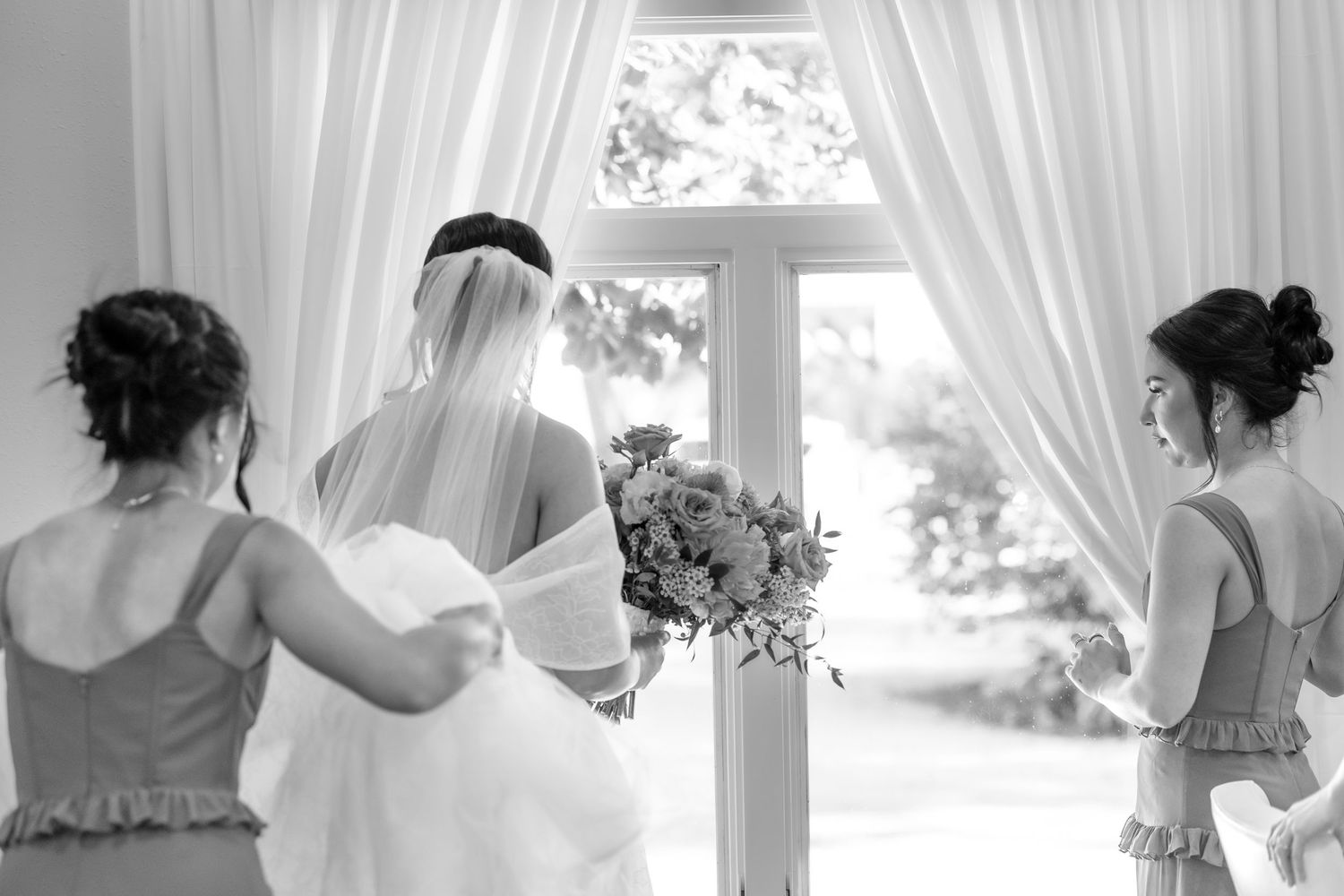 Black and white photo of bride holding bouquet with two bridesmaids adjusting her veil near draped window.