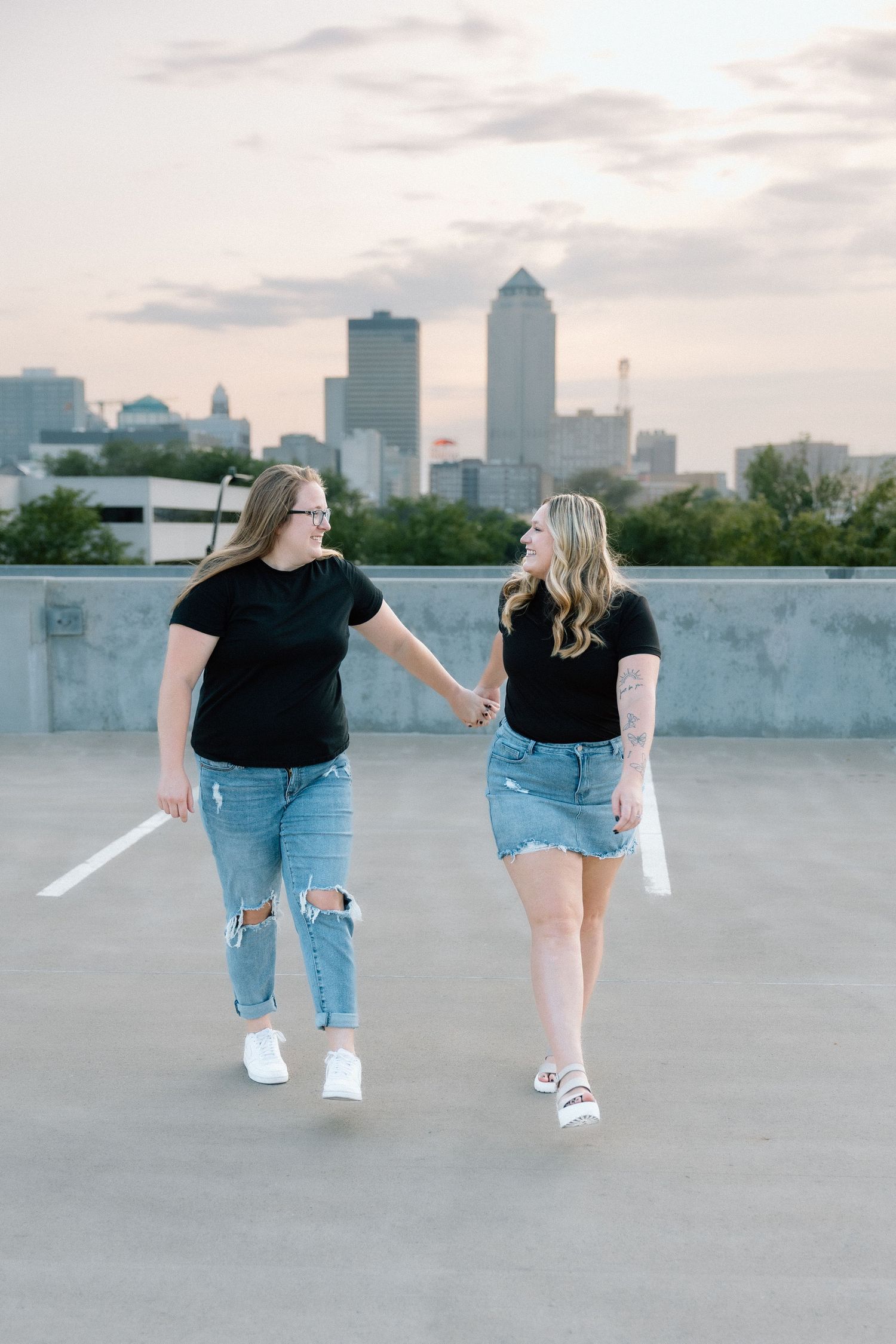Two women holding hands in matching black tees and denim on a rooftop with a city skyline at sunset.