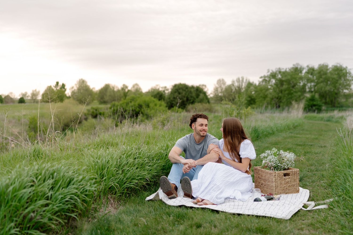Couple sitting on a blanket in a green field having a romantic picnic with a flower basket at sunset.