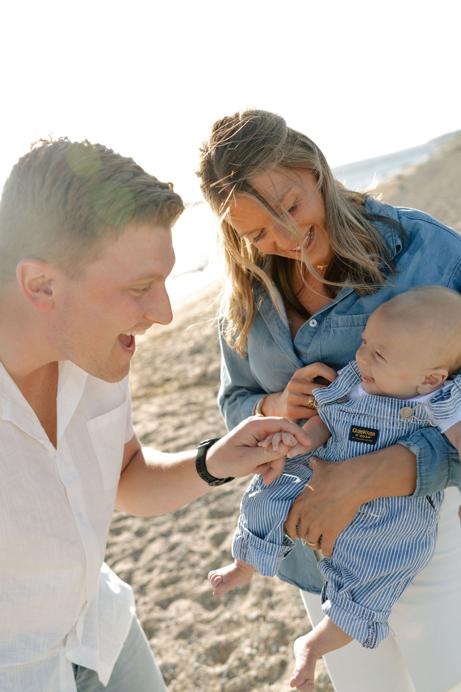 Young family with baby enjoying a sunny beach day, parents smiling at infant in blue striped outfit.