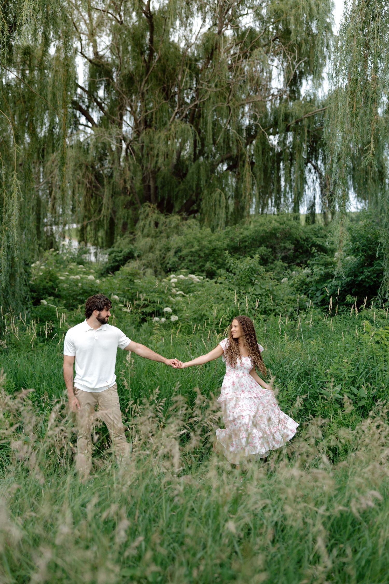 A couple holds hands while walking through a lush green meadow beneath a weeping willow tree.