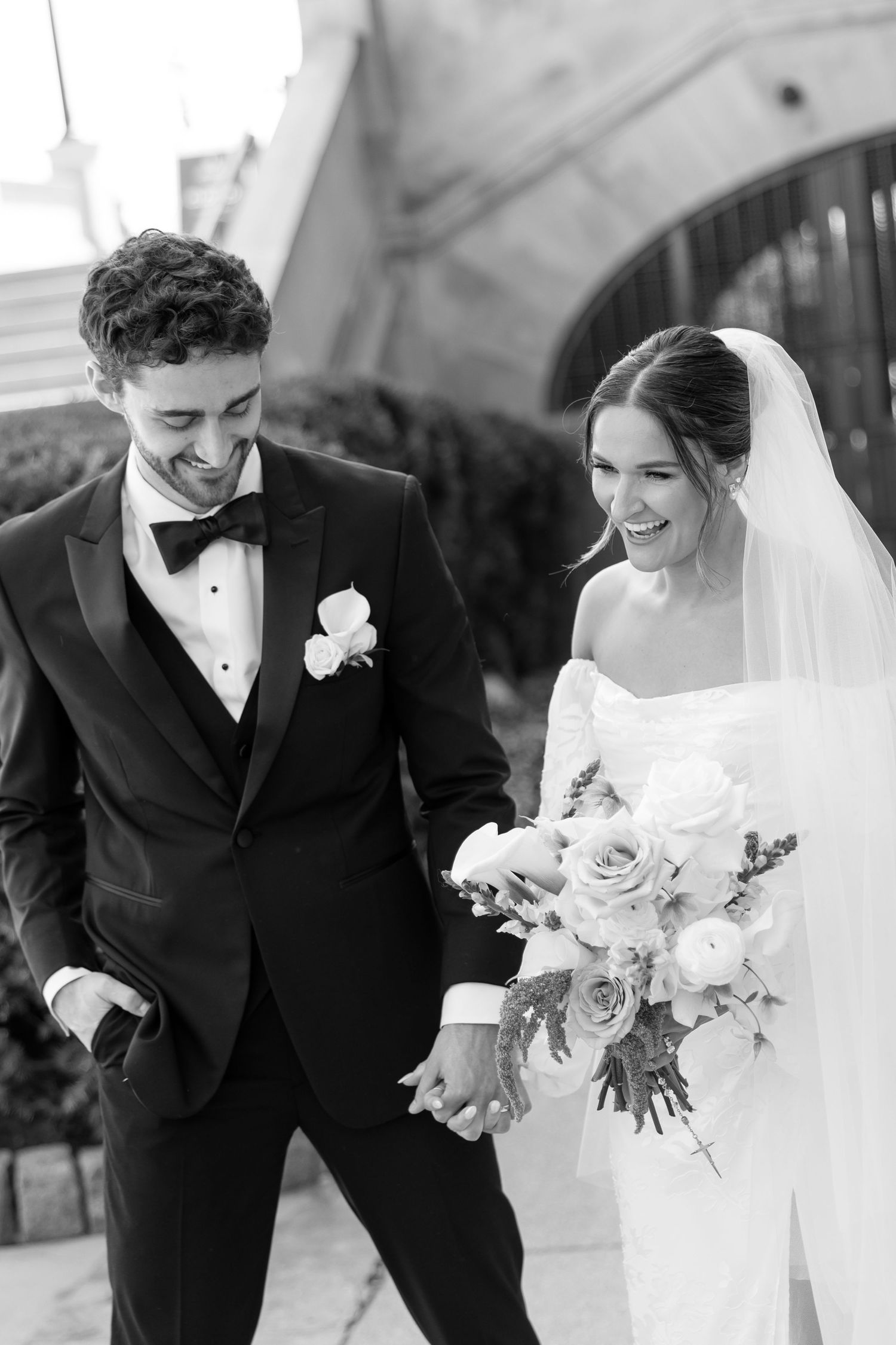 Black and white photo of laughing bride and groom holding hands outside their wedding venue.