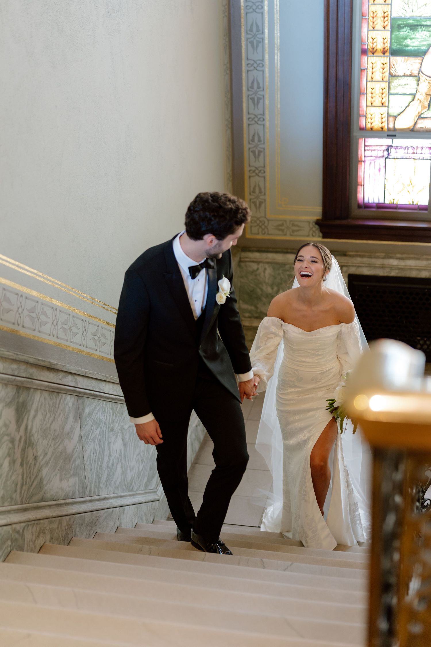 Bride and groom laughing while holding hands on marble staircase, bride in off-shoulder gown, groom in black tuxedo.