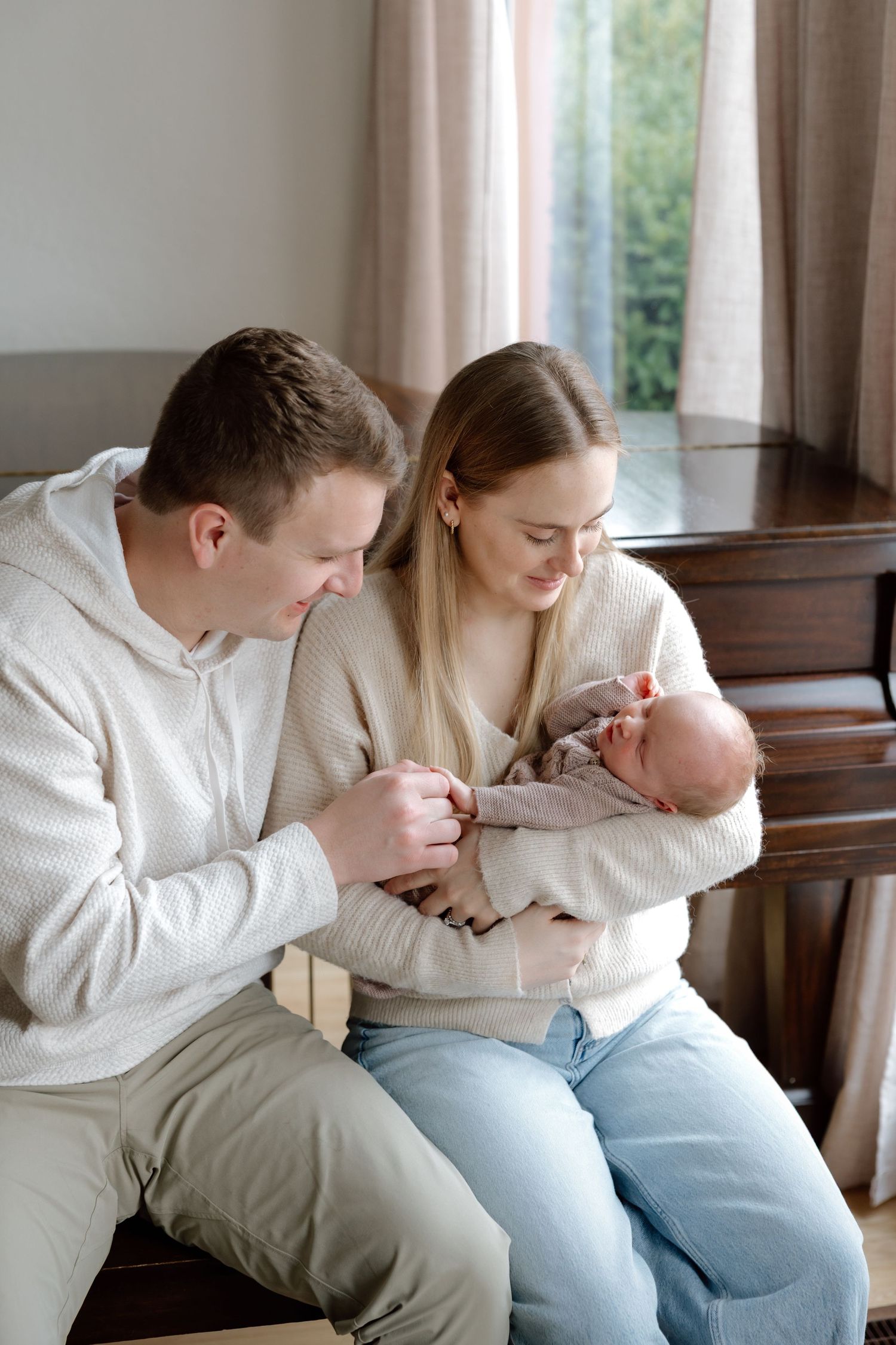 Young parents sit together near a window, smiling as they admire their sleeping newborn baby in a cozy home.