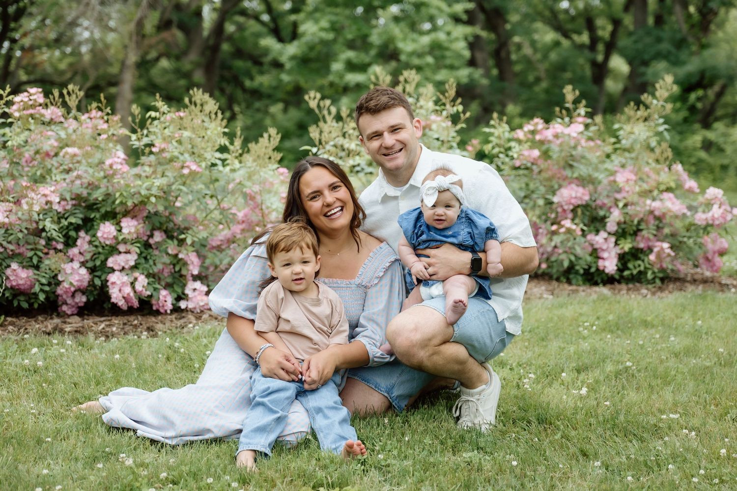 Smiling family of four poses on grass in front of pink flowering bushes with two young children.
