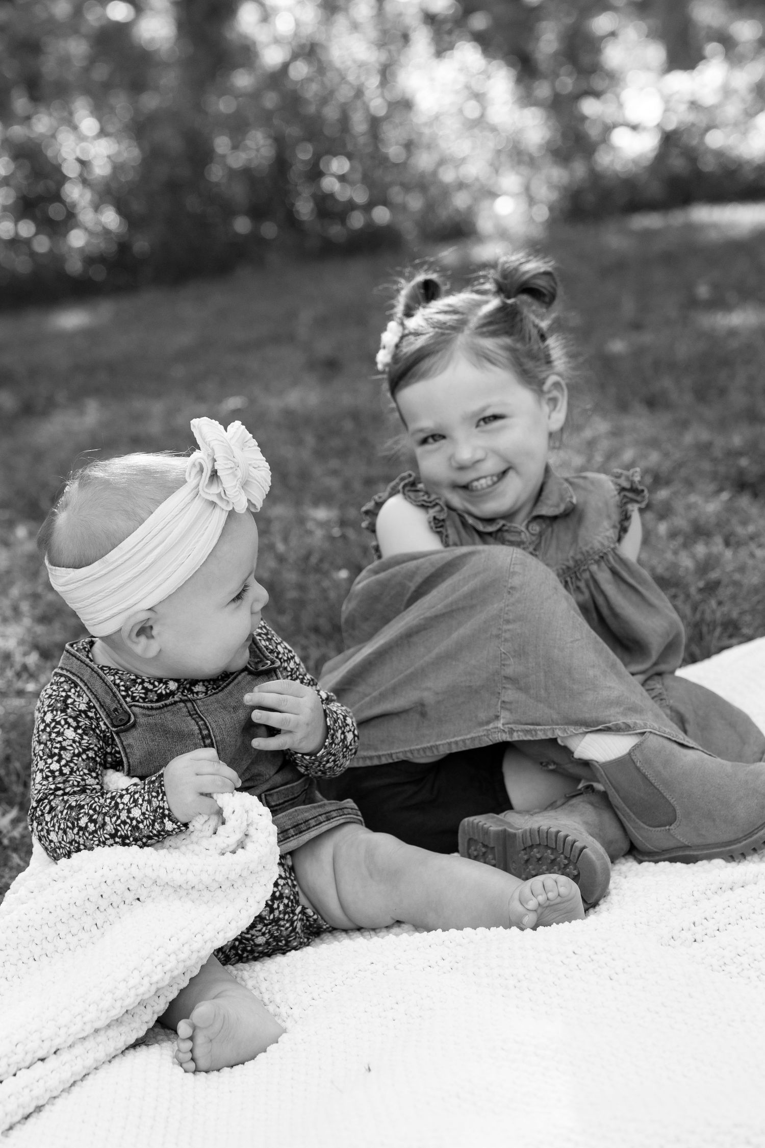 Black and white photo of two young sisters sitting together on a blanket in a park, smiling.