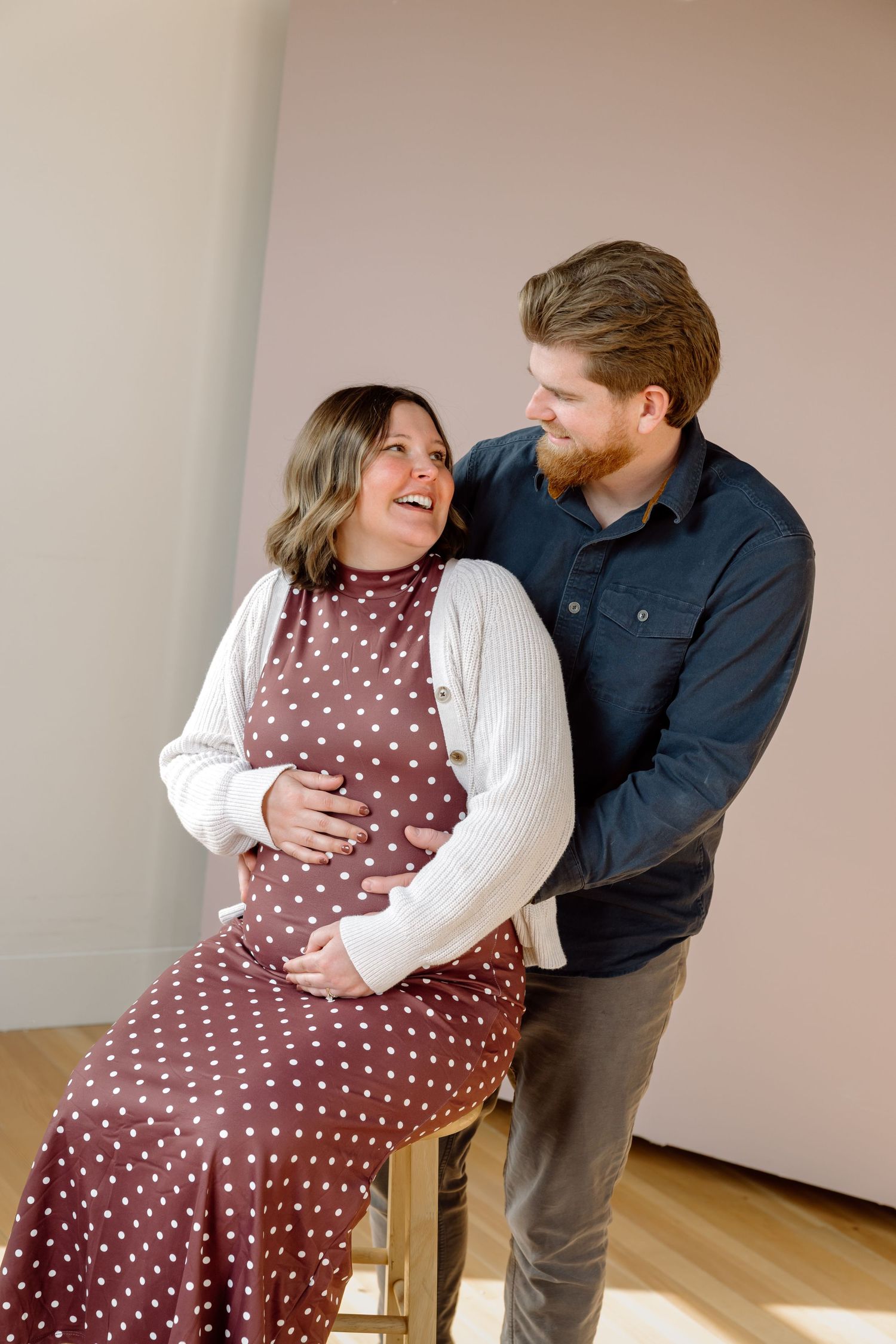 A laughing pregnant woman in a polka dot dress sits as her bearded partner stands beside her smiling.