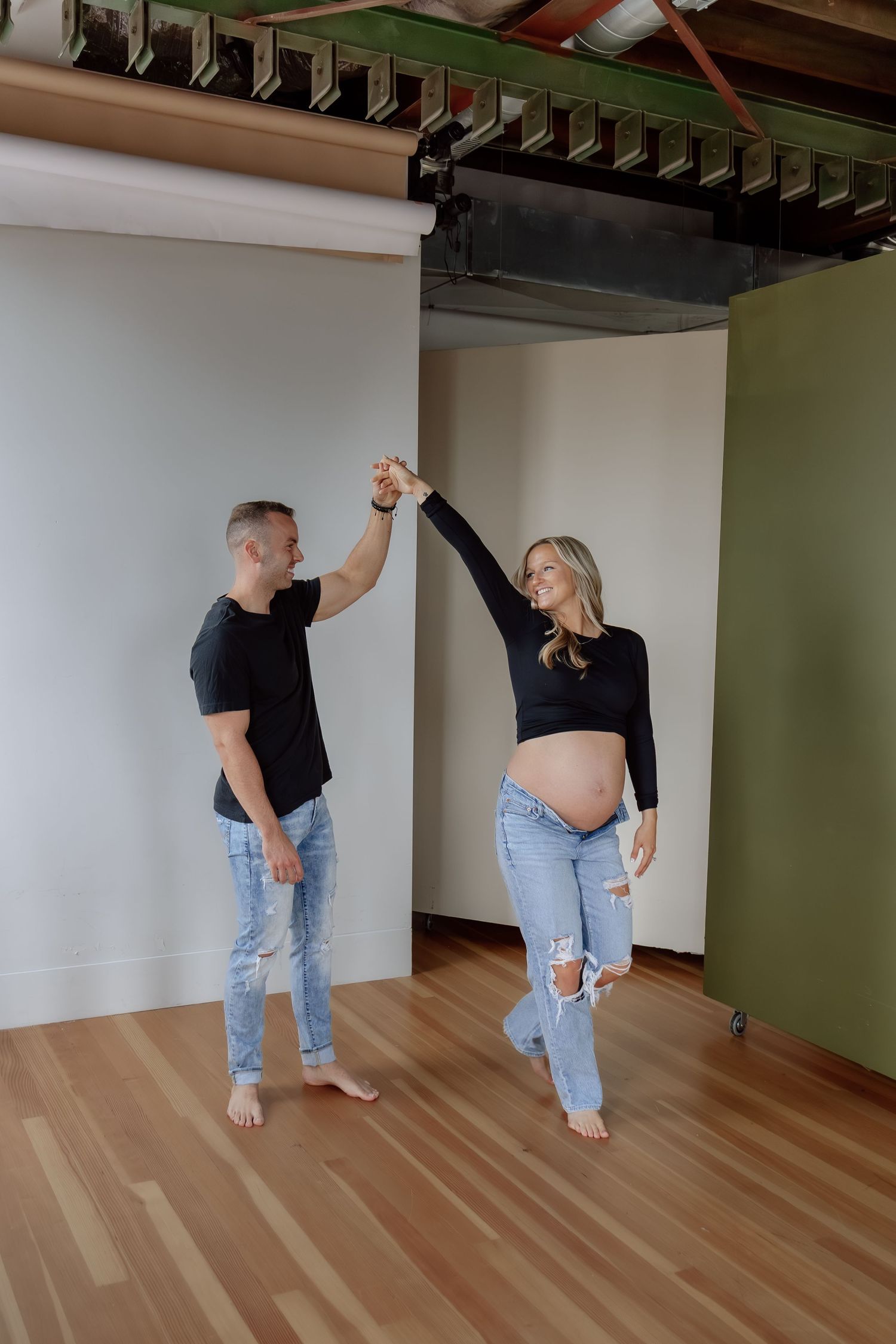 Pregnant couple dancing and holding hands in a bright studio with exposed ceiling beams.